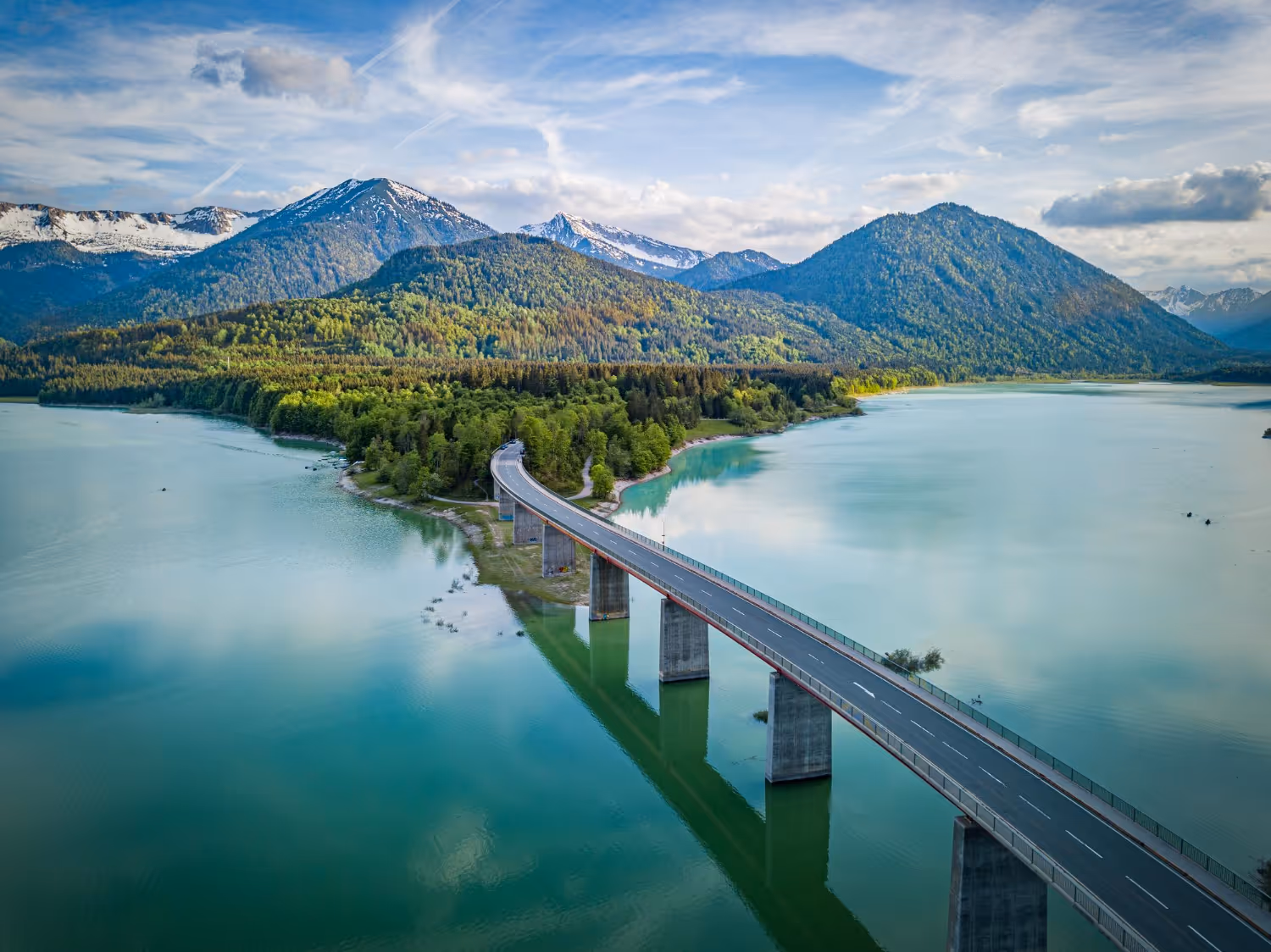 Long pont traversant un lac calme, entouré de collines boisées verdoyantes et de montagnes enneigées, sous un ciel partiellement nuageux.