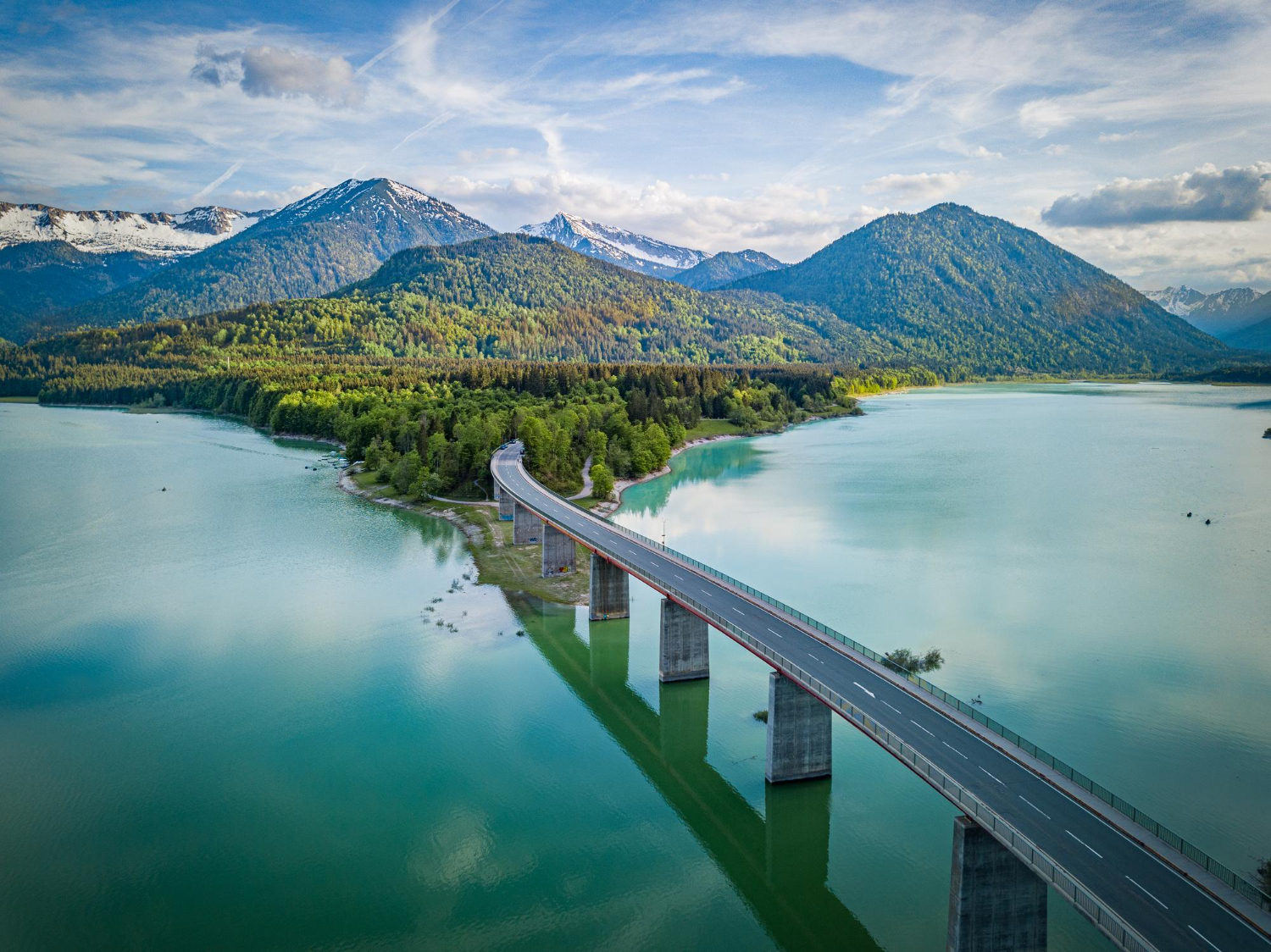 Long pont traversant un lac calme, entouré de collines boisées verdoyantes et de montagnes enneigées, sous un ciel partiellement nuageux.