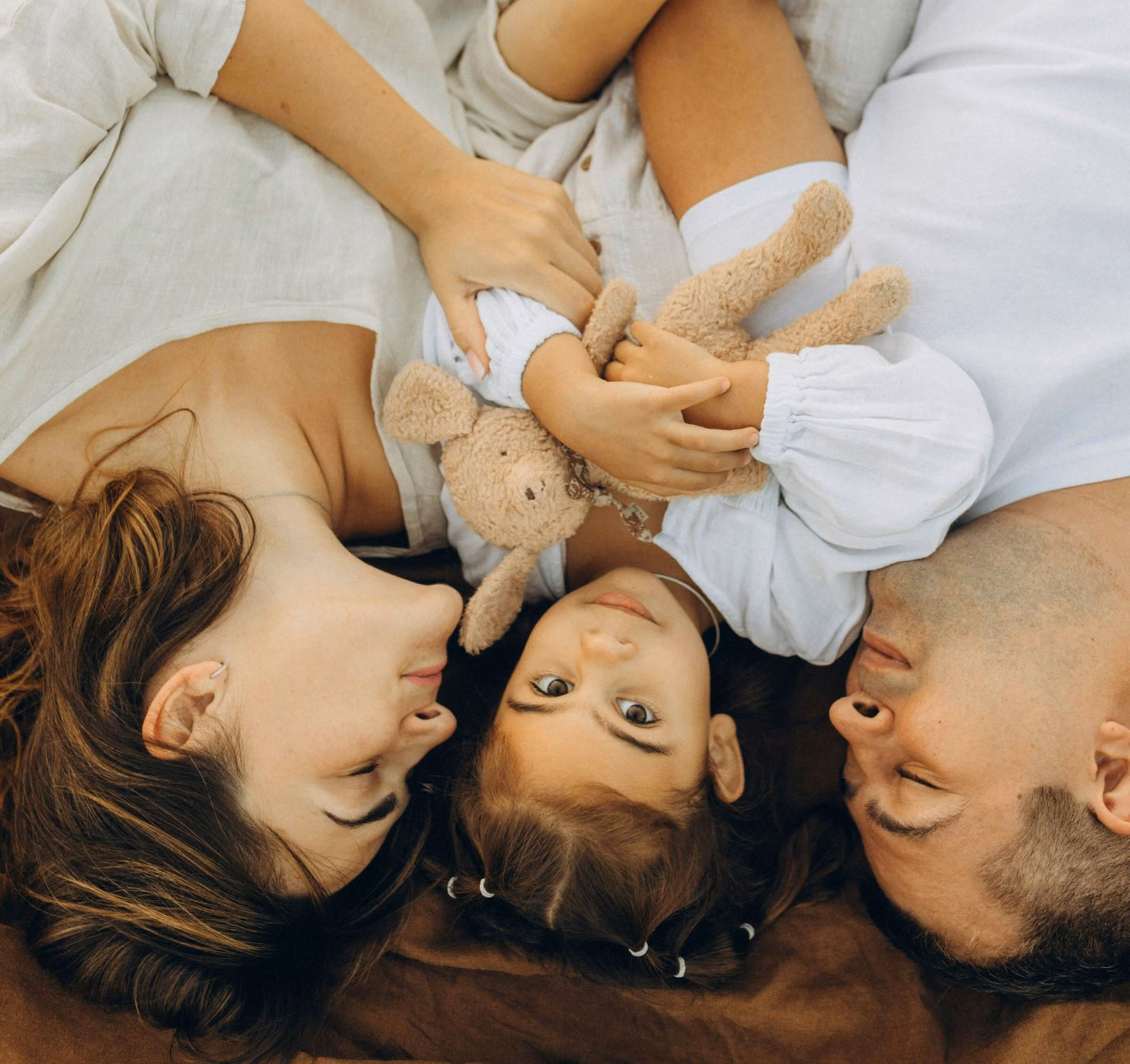 Parents lying on a blanket facing their daughter holding a plush bunny toy between them.