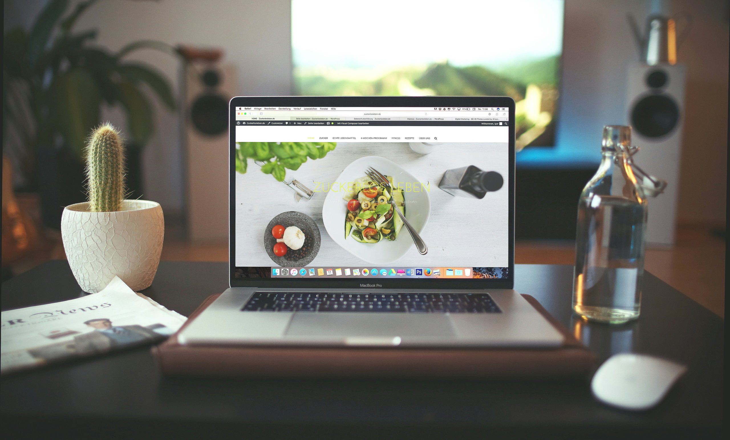 Laptop on a desk displaying a website with food images, next to a cactus in a pot, a newspaper, a glass bottle, and a white mouse.