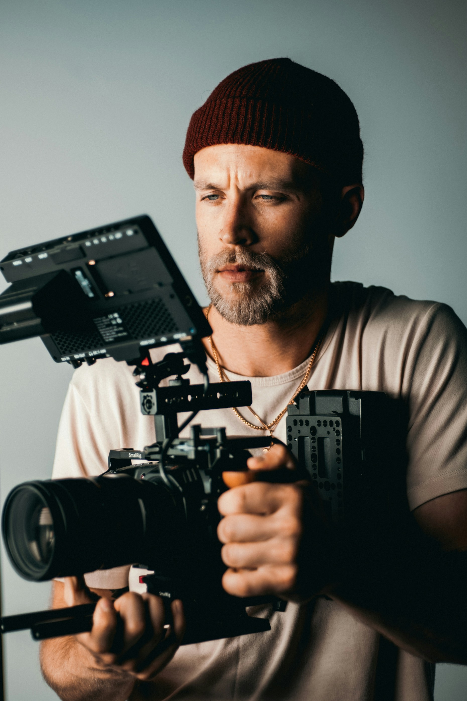 Bearded man wearing a maroon beanie and gold necklaces holding a professional video camera rig with a large lens and monitor.
