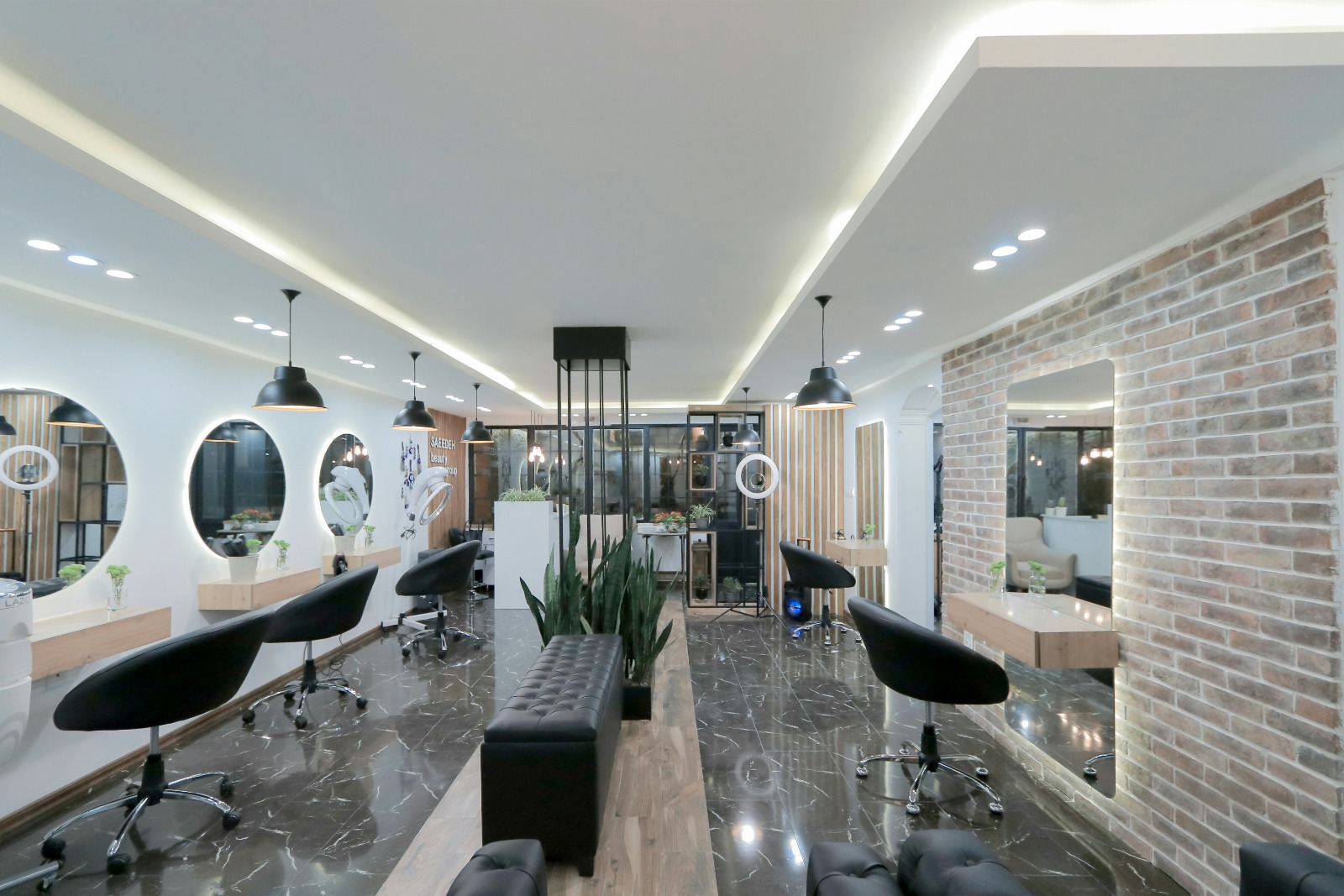 Interior of a modern beauty salon with black swivel chairs, large mirrors, pendant lights, plants, and marble flooring.