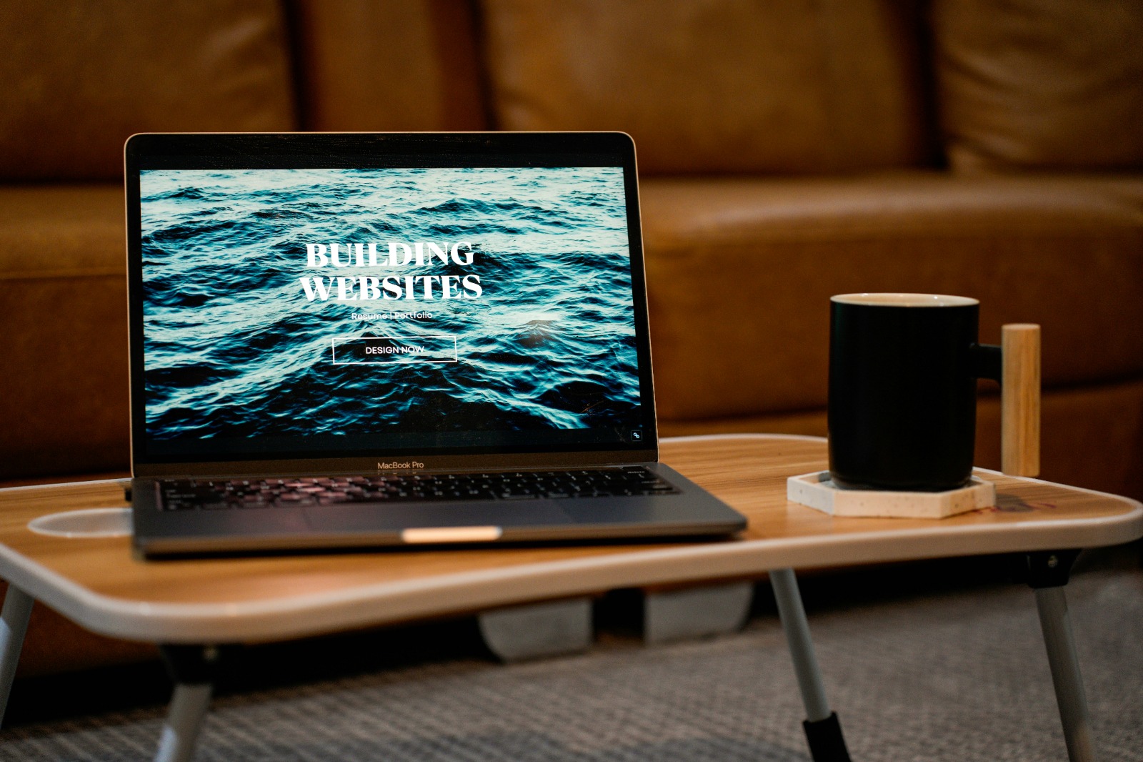 MacBook Pro laptop on a wooden tray table showing a website with the text 'Building Websites' over an ocean waves background, next to a black mug on a coaster.