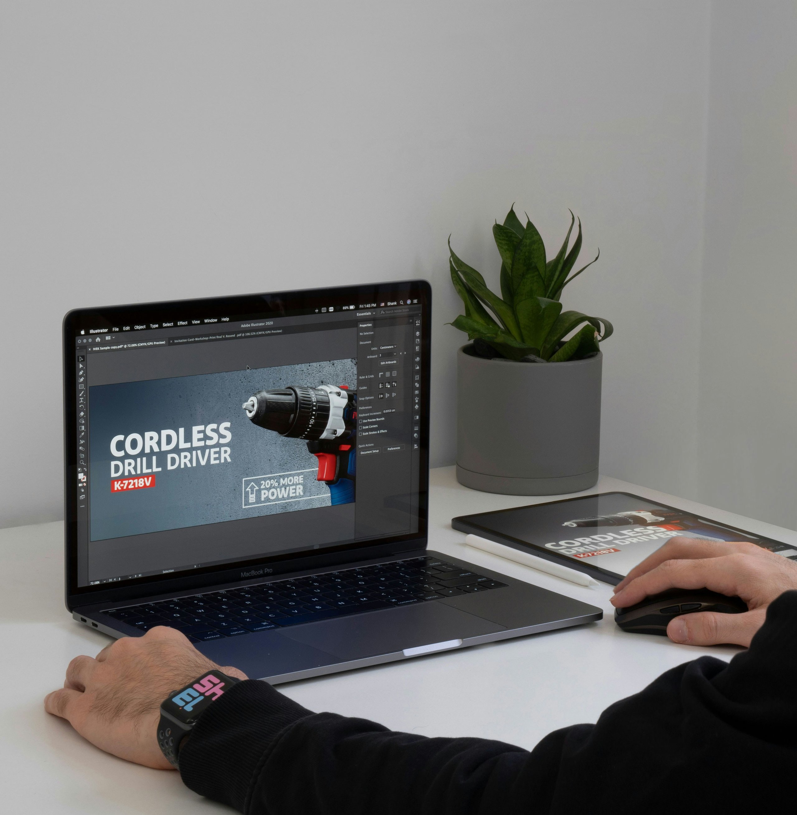 Person working on a MacBook Pro editing a cordless drill driver advertisement, with a potted plant and tablet on a white desk.
