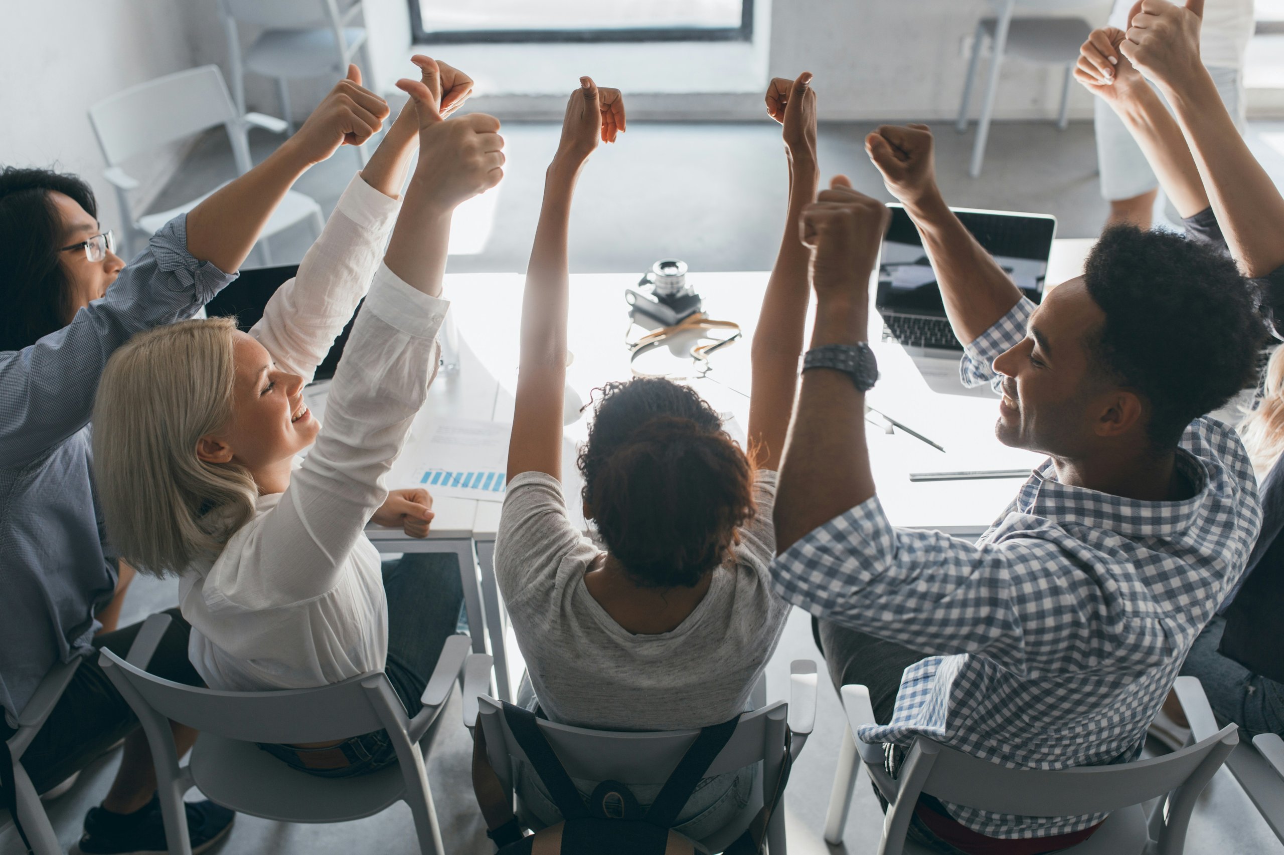 Diverse group of coworkers seated around a table, raising their fists and thumbs in a gesture of celebration and unity.