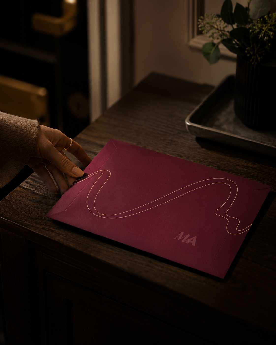 Une main saisit une enveloppe framboise scellée, posée sur une console le soir