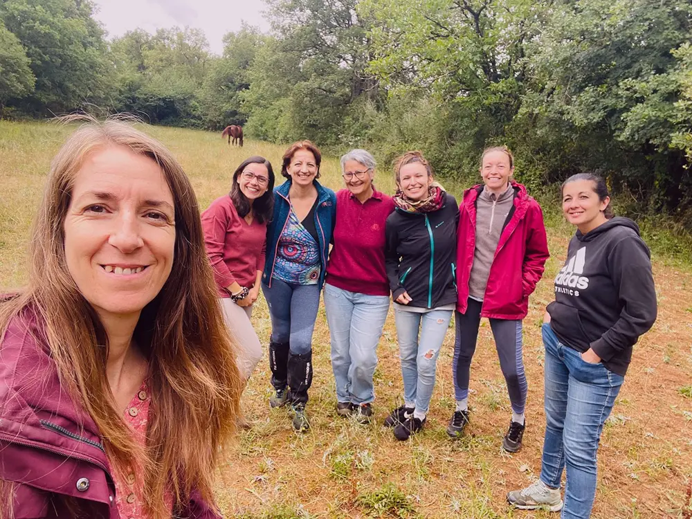 Barbara Edelmann et 6 élèves de la formation Coach Equin Holistique, souriantes debout dans un champ herbeux avec des arbres en arrière-plan et un cheval au loin.