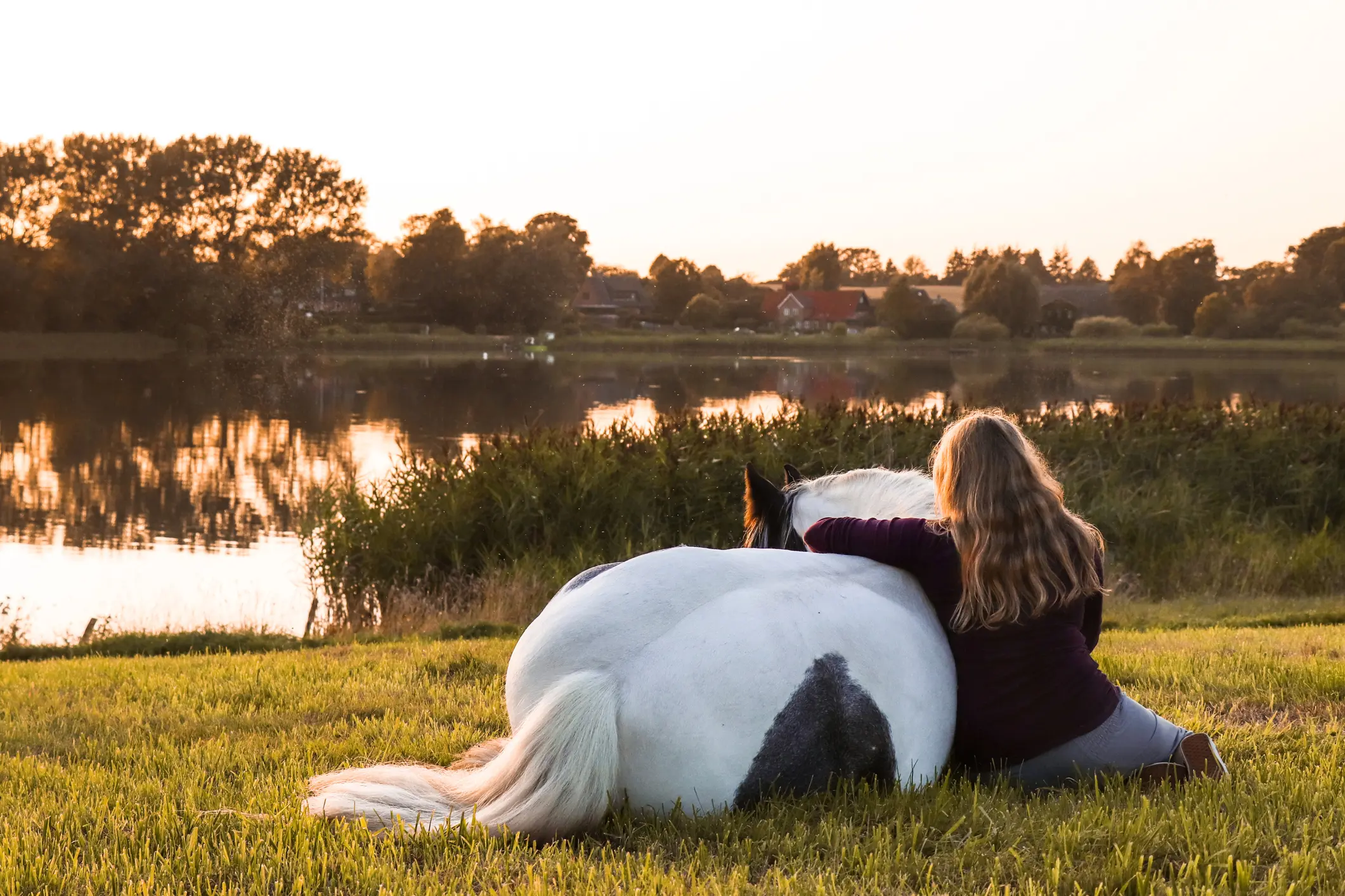 Une femme aux cheveux longs assise dans l’herbe avec un cheval blanc et noir allongé près d’un lac au coucher du soleil.
