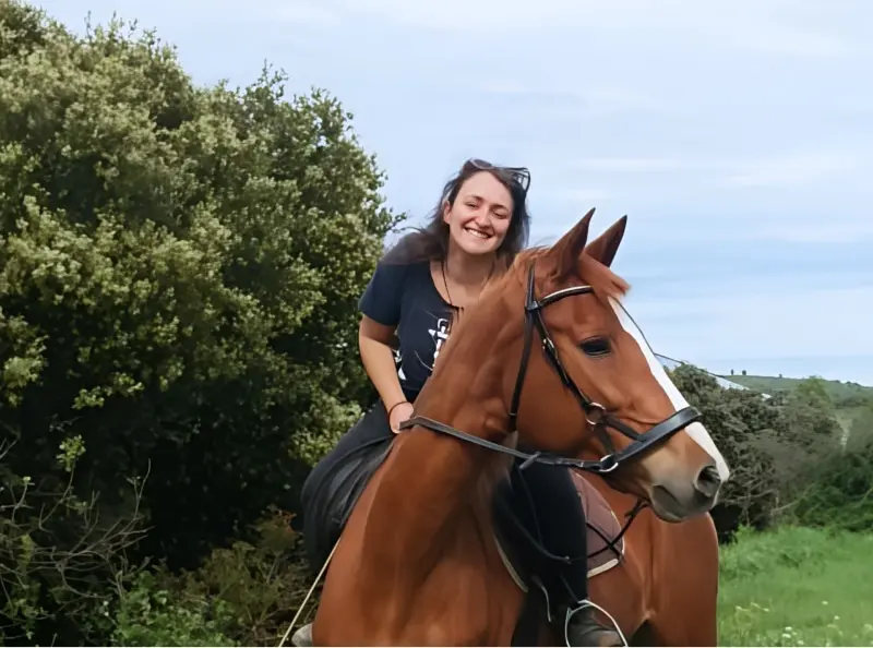 Femme souriante sur un cheval marron avec une marque blanche sur le visage, dans un paysage verdoyant avec des arbustes.
