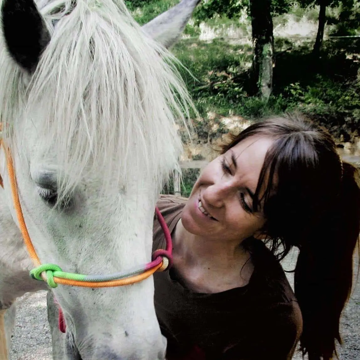 Une femme souriante en train de caresser un cheval blanc portant un licol multicolore, en extérieur avec des arbres en arrière-plan.