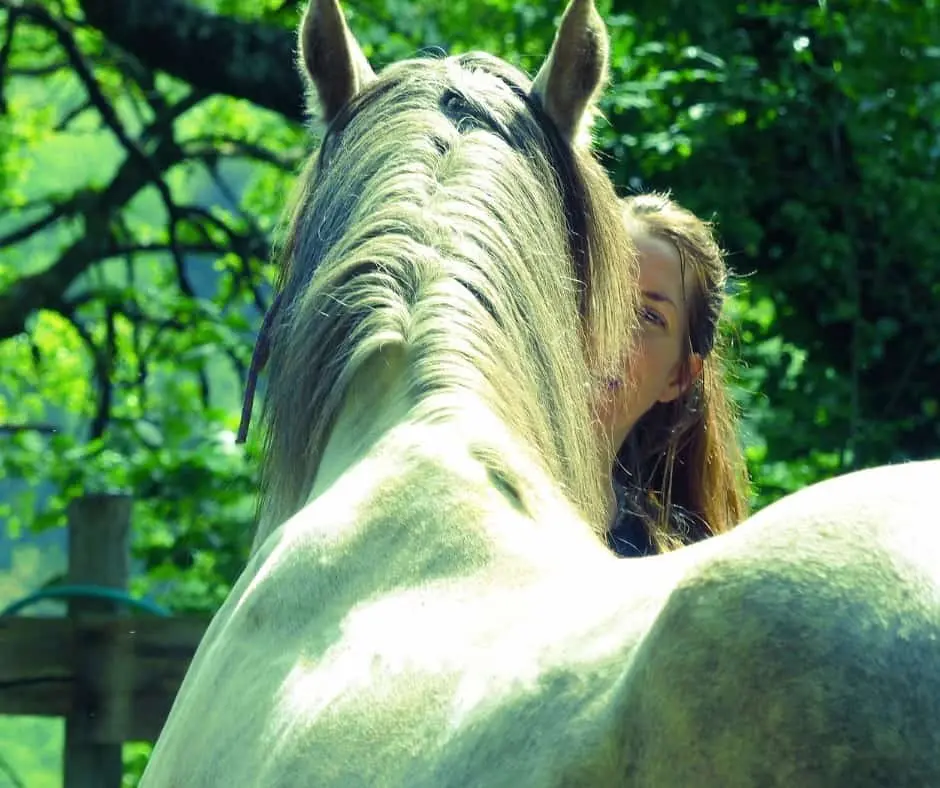 Barbara Edelmann se tient derrière un cheval gris clair dans un cadre naturel avec des arbres verts en arrière-plan.