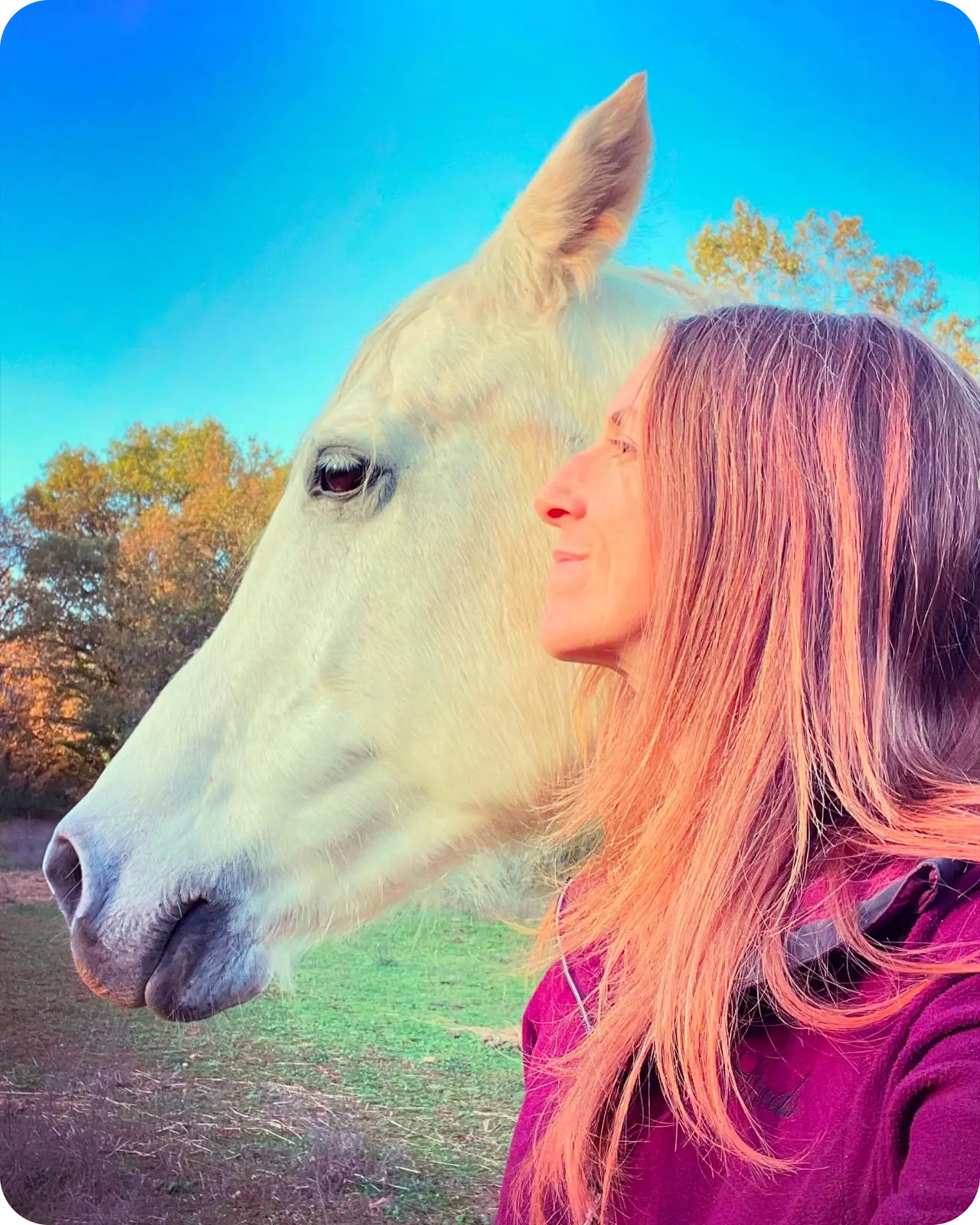 Barbara Edelmann pose à côté d’un cheval blanc dans un paysage herbeux sous un ciel bleu.