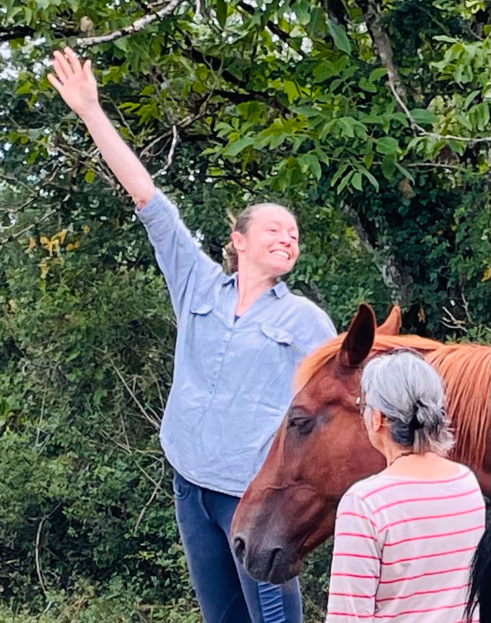 Une femme souriante lève un bras dans un geste joyeux à côté d'un cheval marron et d'une autre personne aux cheveux gris portant un t-shirt rayé.