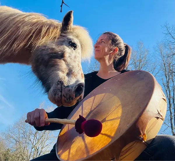 Une femme jouant d'un grand tambour à main près d'un poney blanc sous un ciel bleu clair.