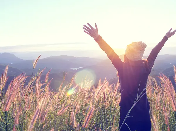 Femme levant les bras face au soleil couchant dans un champ d'herbes hautes avec des montagnes en arrière-plan.