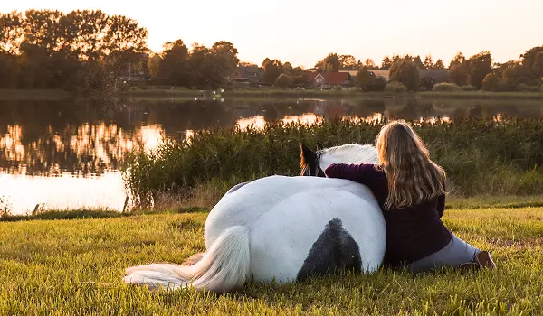 Une femme assise sur l'herbe au bord d'un lac embrassant un cheval blanc tacheté de noir allongé à côté d'elle au coucher du soleil.