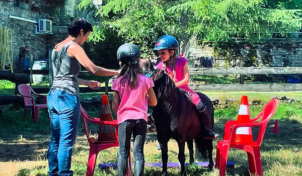 Une fillette casquée en cours d'équitation sur un poney noir avec deux femmes autour dans un enclos herbeux avec chaises rouges et cônes de signalisation.