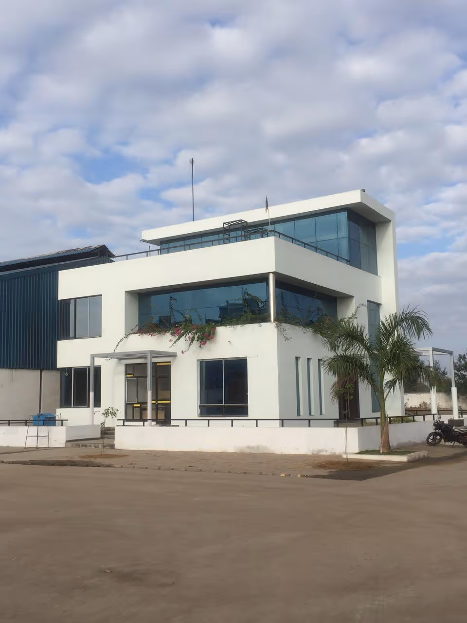 Modern two-story white office building with large blue-tinted windows, plants on the balcony, and a palm tree nearby under a partly cloudy sky.