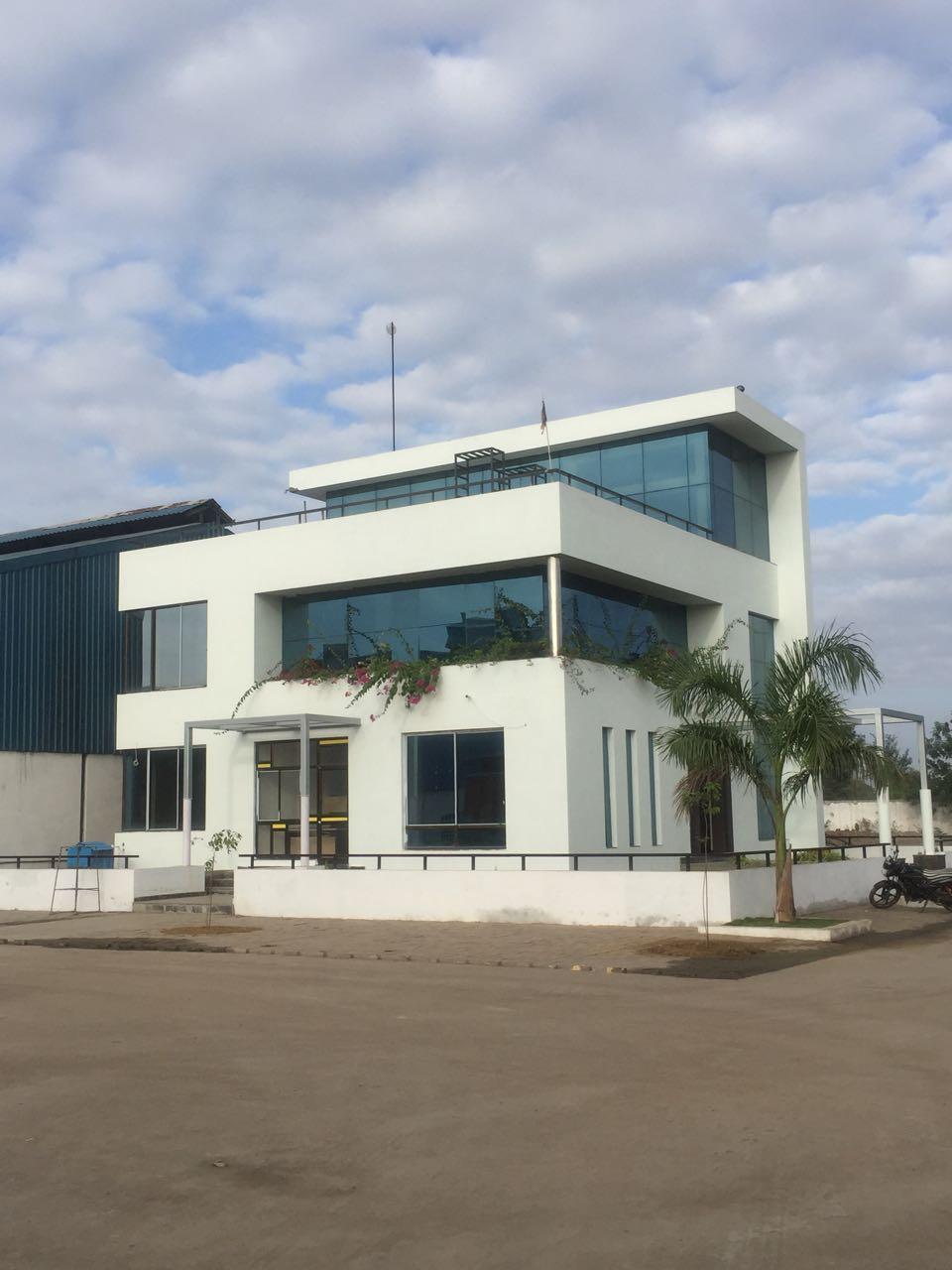 Modern two-story white office building with large blue-tinted windows, plants on the balcony, and a palm tree nearby under a partly cloudy sky.