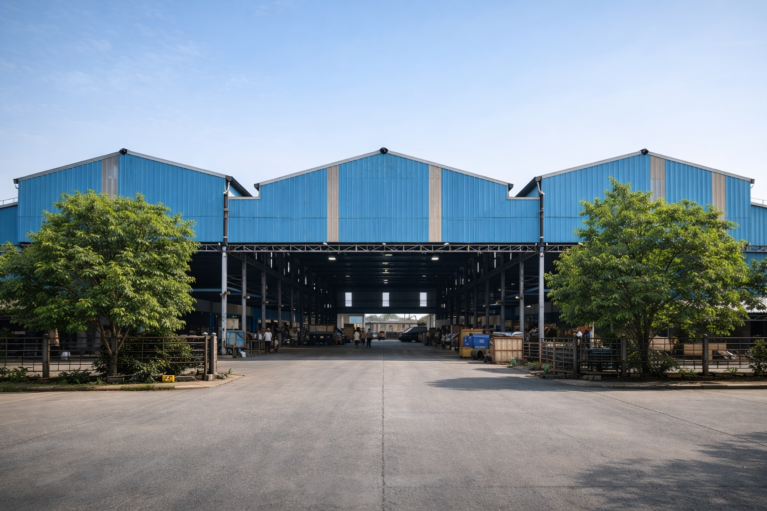 Large industrial warehouse with blue metal walls and open entrance, flanked by green trees on both sides.