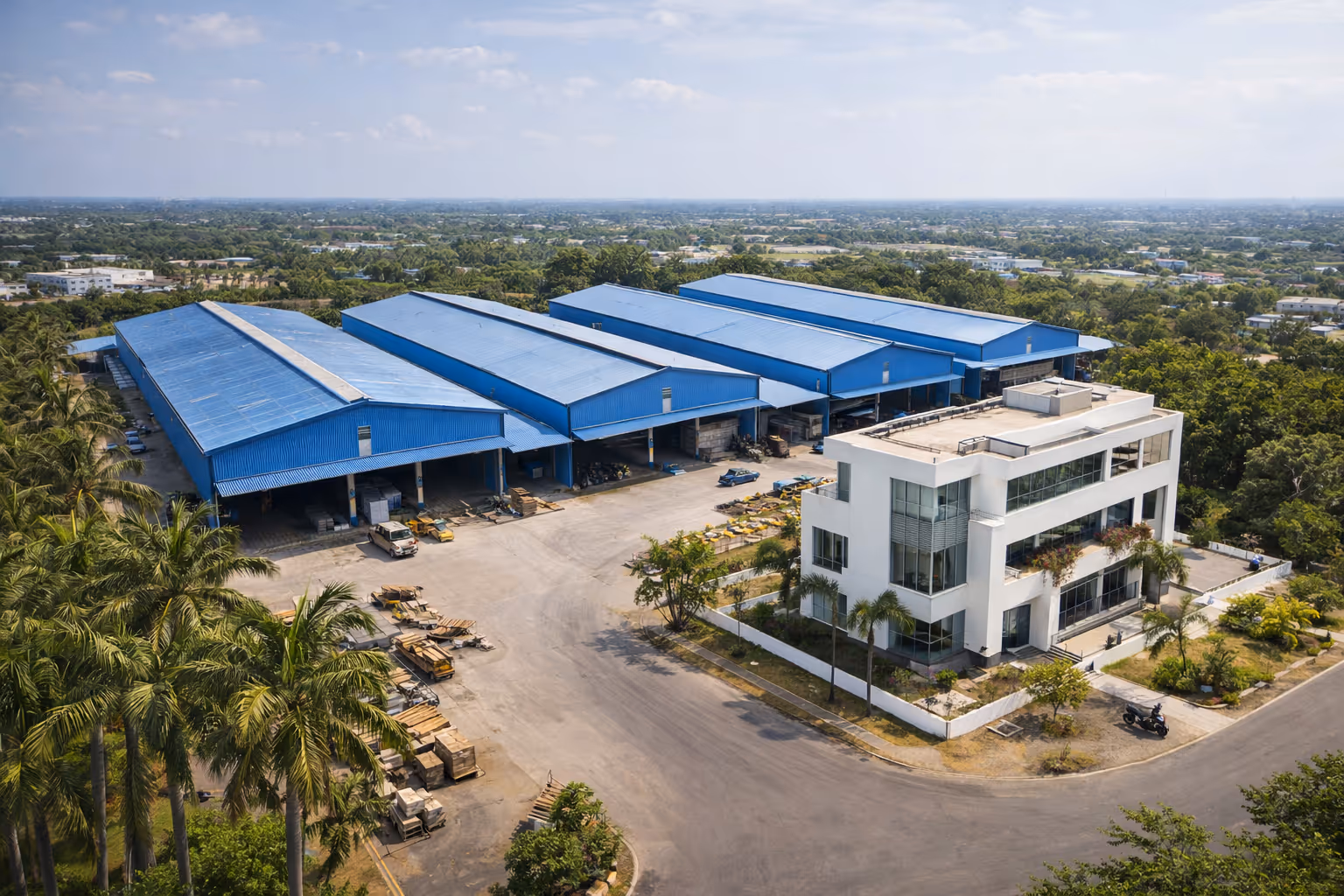 Aerial view of an industrial complex with four large blue warehouses and a modern white office building surrounded by trees and open paved areas.