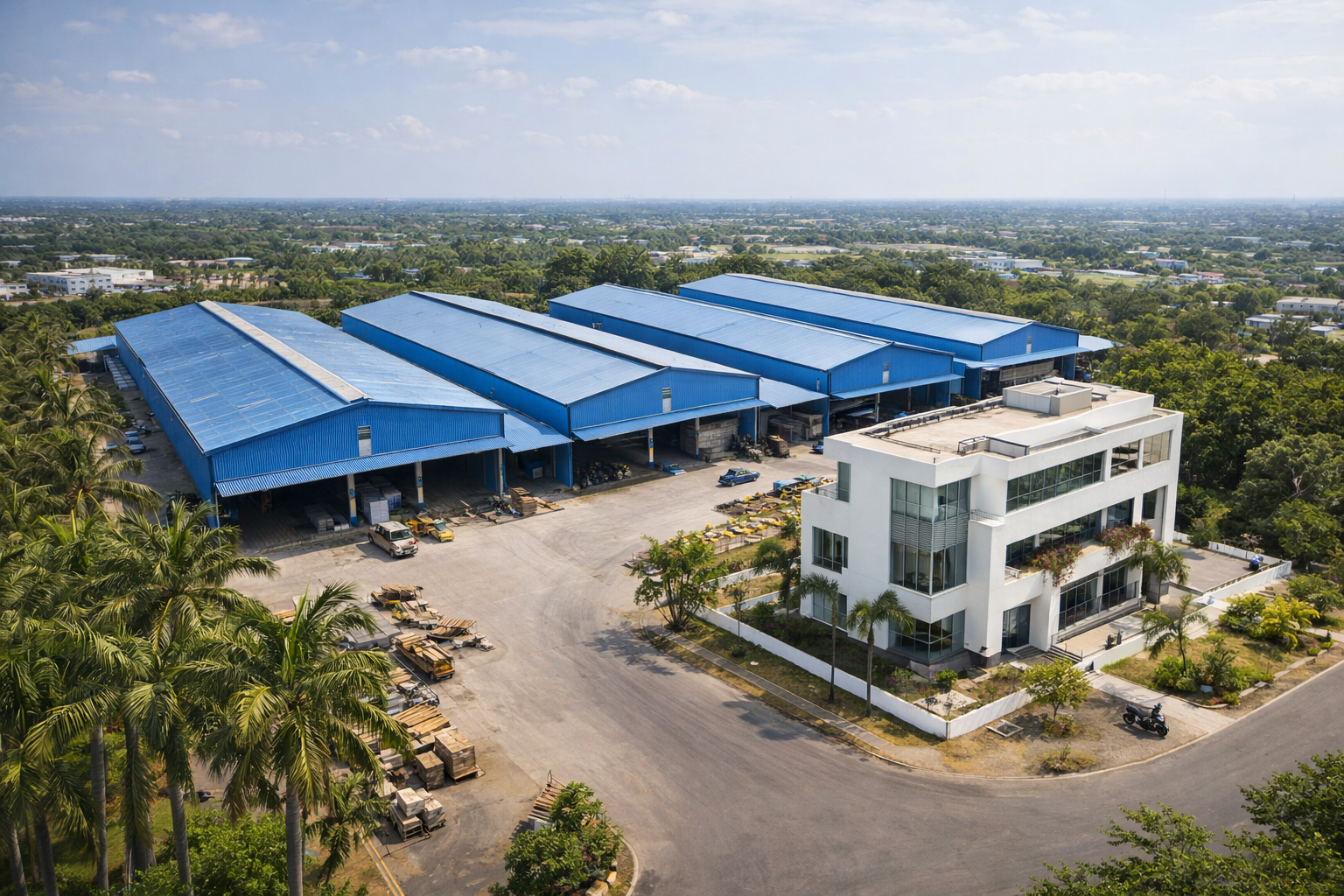 Aerial view of an industrial complex with four large blue warehouses and a modern white office building surrounded by trees and open paved areas.