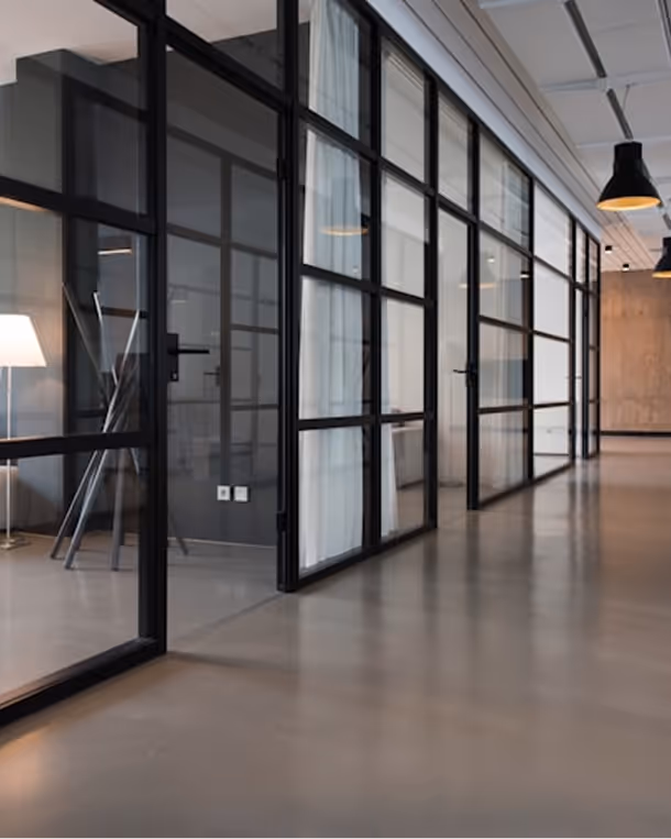 Modern office hallway with glass-walled conference rooms featuring black metal frames and a polished concrete floor.