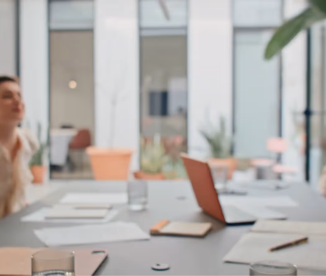 Blurred office meeting room with a laptop, notebooks, papers, glasses of water, and a potted plant.