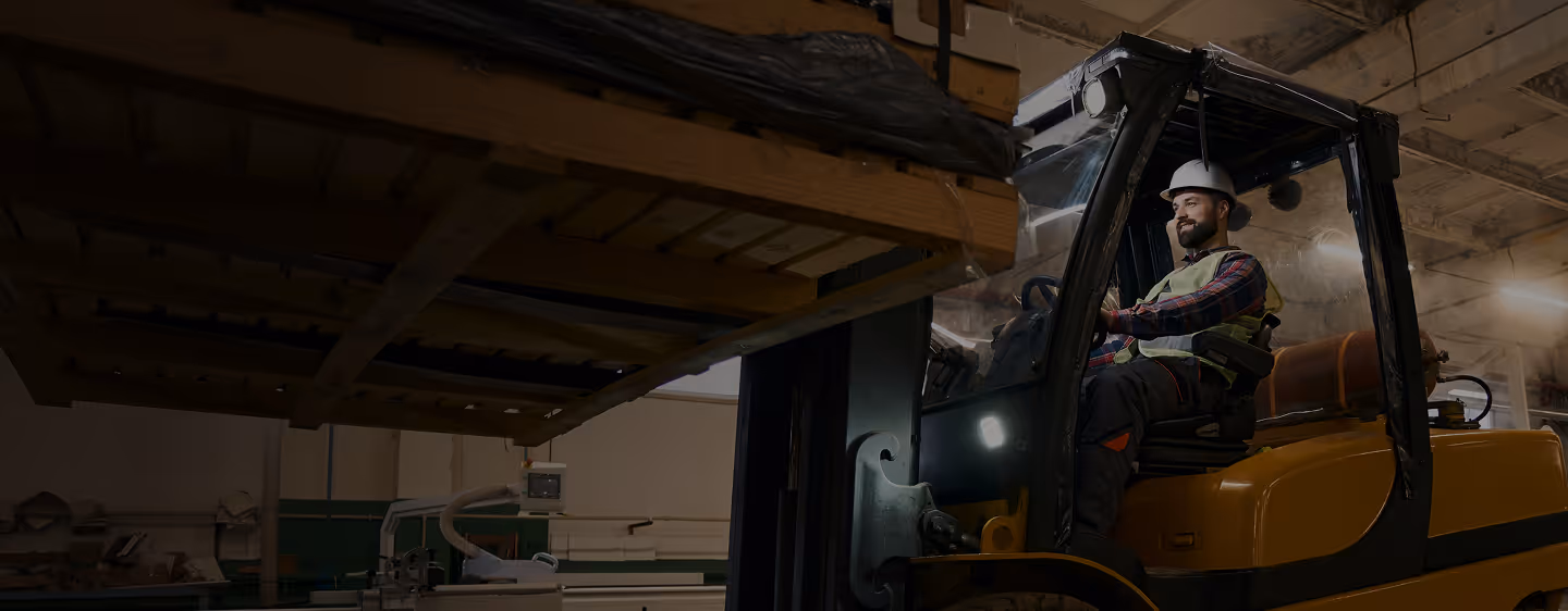 Worker wearing a white hard hat and safety vest operating a yellow forklift inside a warehouse.