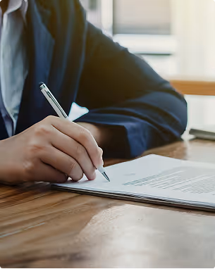 Person in a navy blazer signing a document on a wooden table.