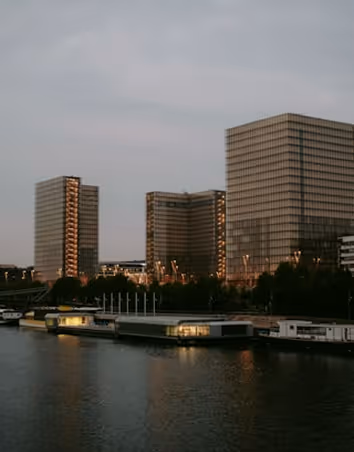 Modern glass office buildings reflecting evening light along a riverside with boats docked in the foreground.