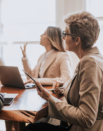 Two professional women in a meeting, one holding a tablet and stylus while the other gestures near a laptop.