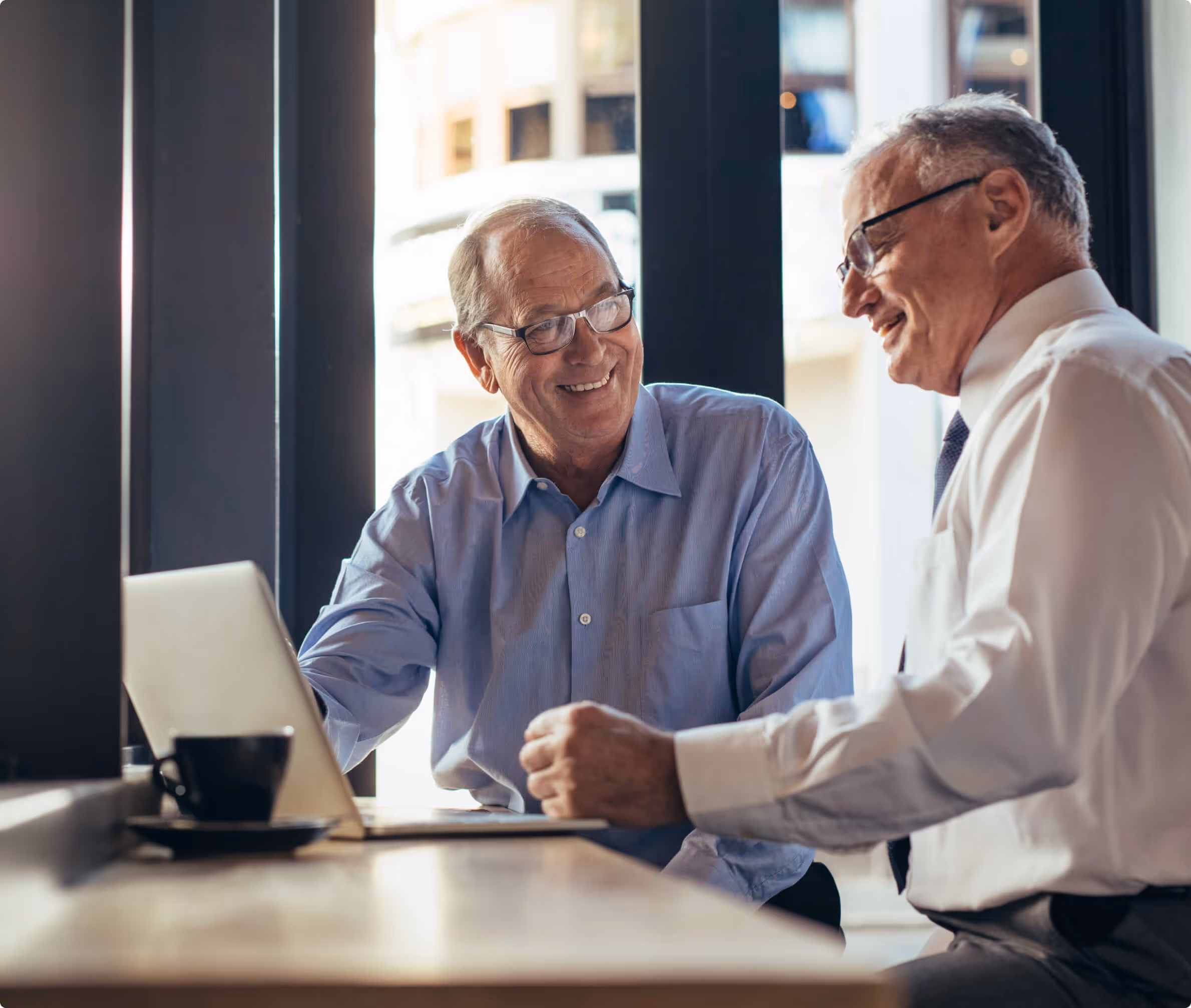 Two elderly men wearing glasses smiling and looking at a laptop while sitting at a table by a window.