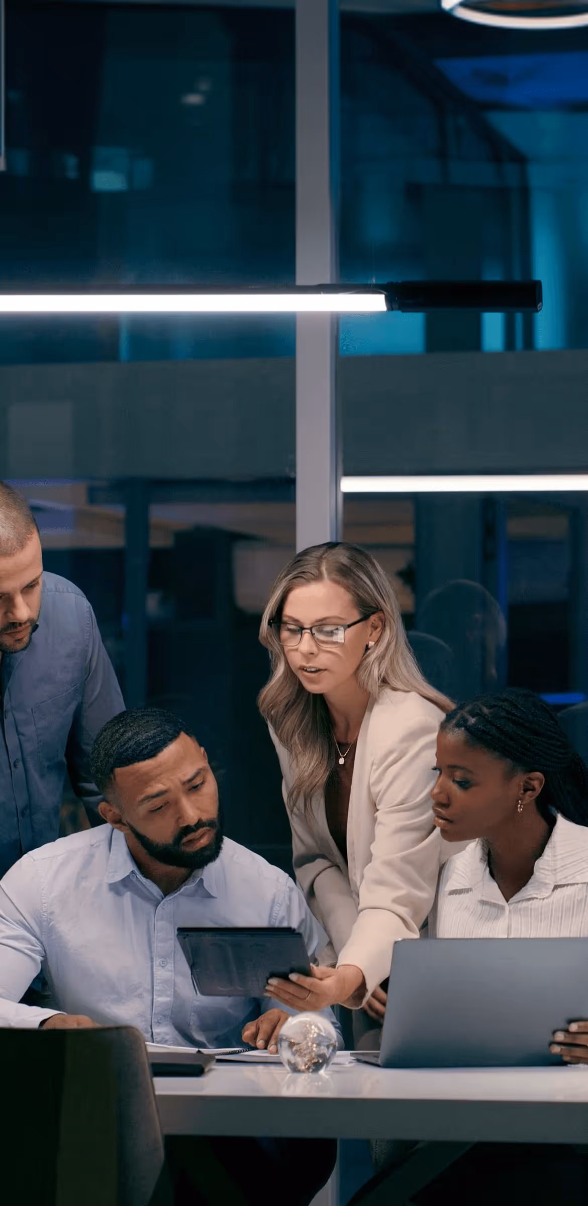 Four diverse colleagues collaborating around a table, reviewing a tablet and laptop in a modern office.
