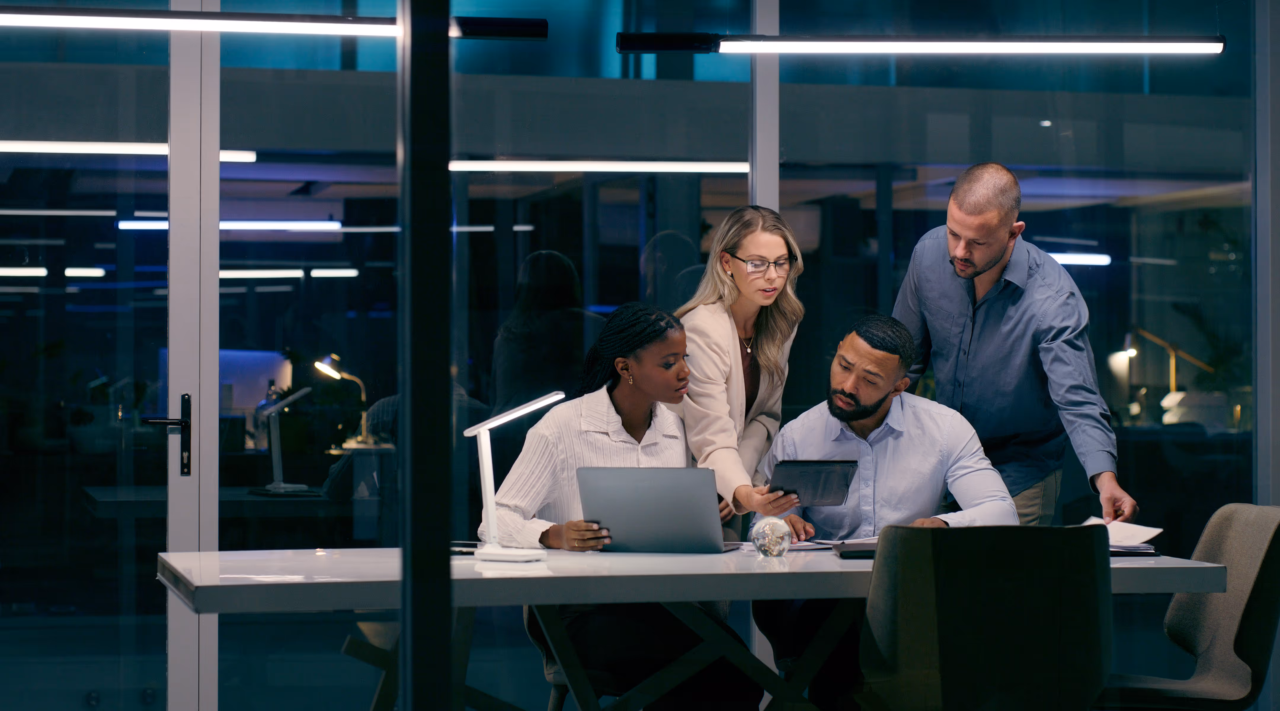 Four colleagues collaborating in a modern office at night, reviewing information on a tablet and laptop around a white table.
