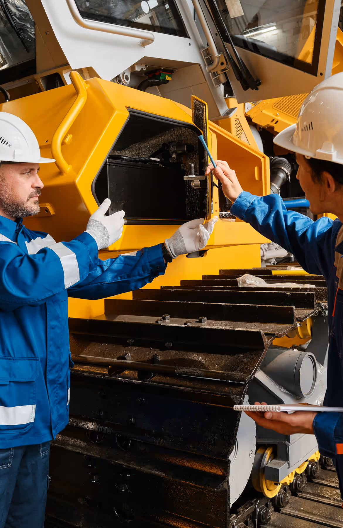 Two workers in safety helmets and blue uniforms inspecting the engine compartment of a large yellow construction vehicle.