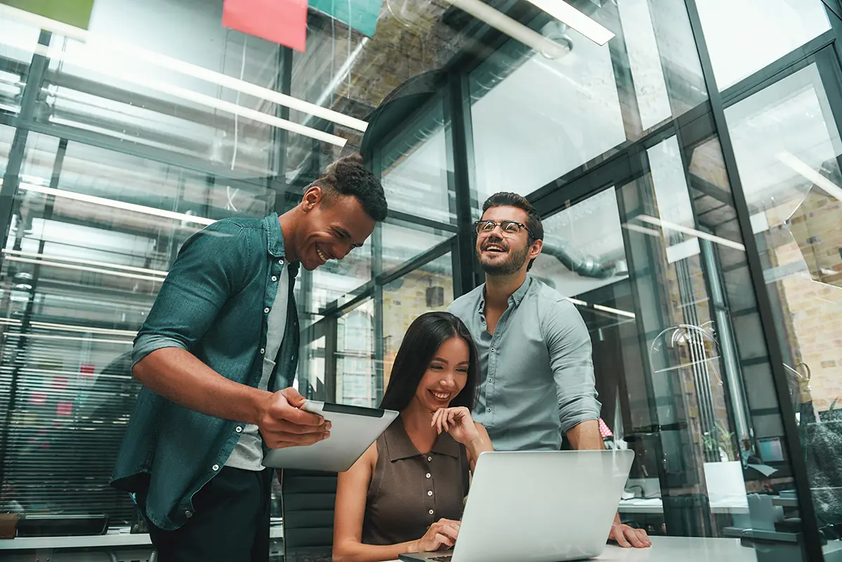 Three young professionals collaborating and smiling while looking at a laptop and tablet in a modern glass office.