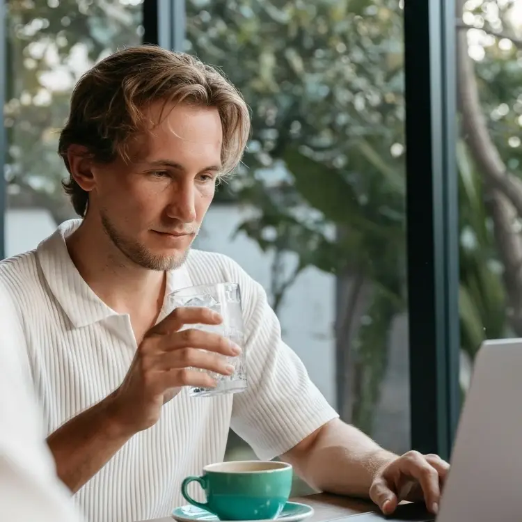 Man in a white shirt sitting at a table, holding a glass of water and working on a laptop in a café with large windows and greenery outside.
patrick zike drinks coffee