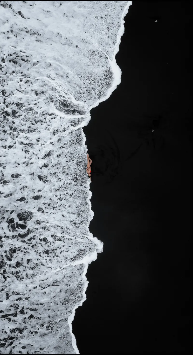 Aerial view of white ocean waves crashing against a person lying on black sand beach.