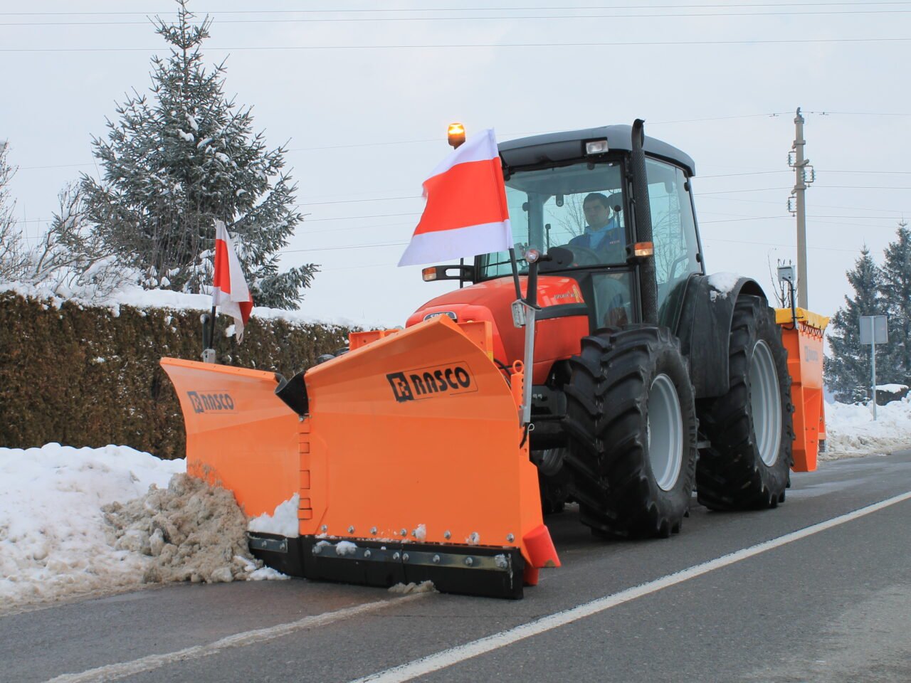 Orange snowplow tractor clearing snow on a road beside a snow-covered hedge and trees.
