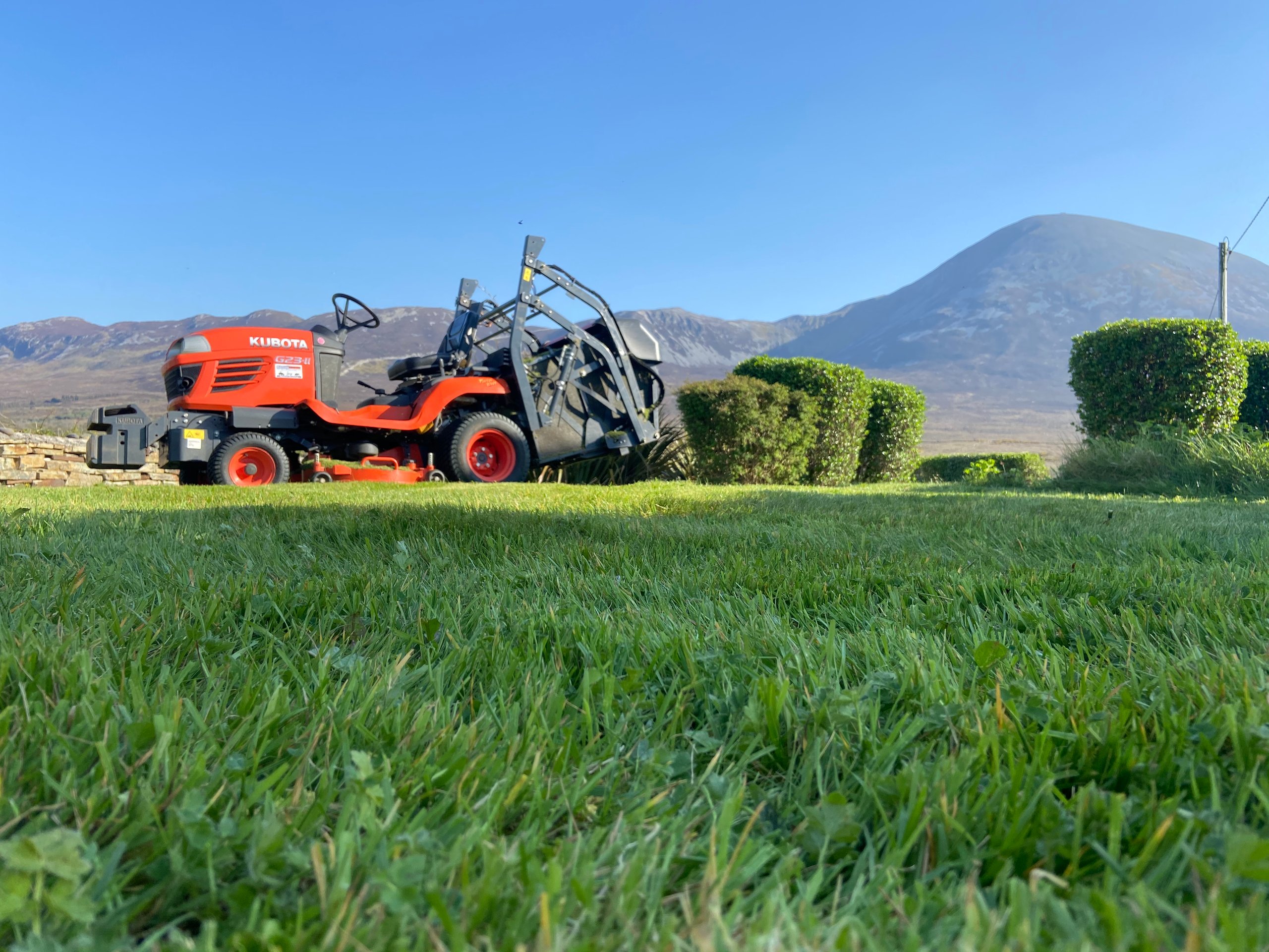 Orange Kubota G23-II lawn mower parked on green grass with trimmed bushes and mountains in the background under a clear blue sky.