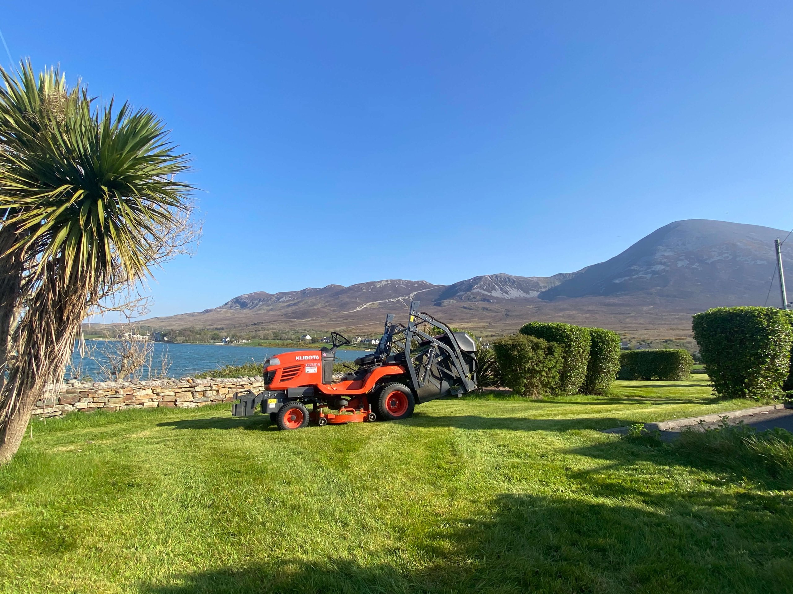 Red Kubota lawn tractor on freshly mowed grass near trimmed hedges, with a lake on left and mountains in the background under a clear blue sky.