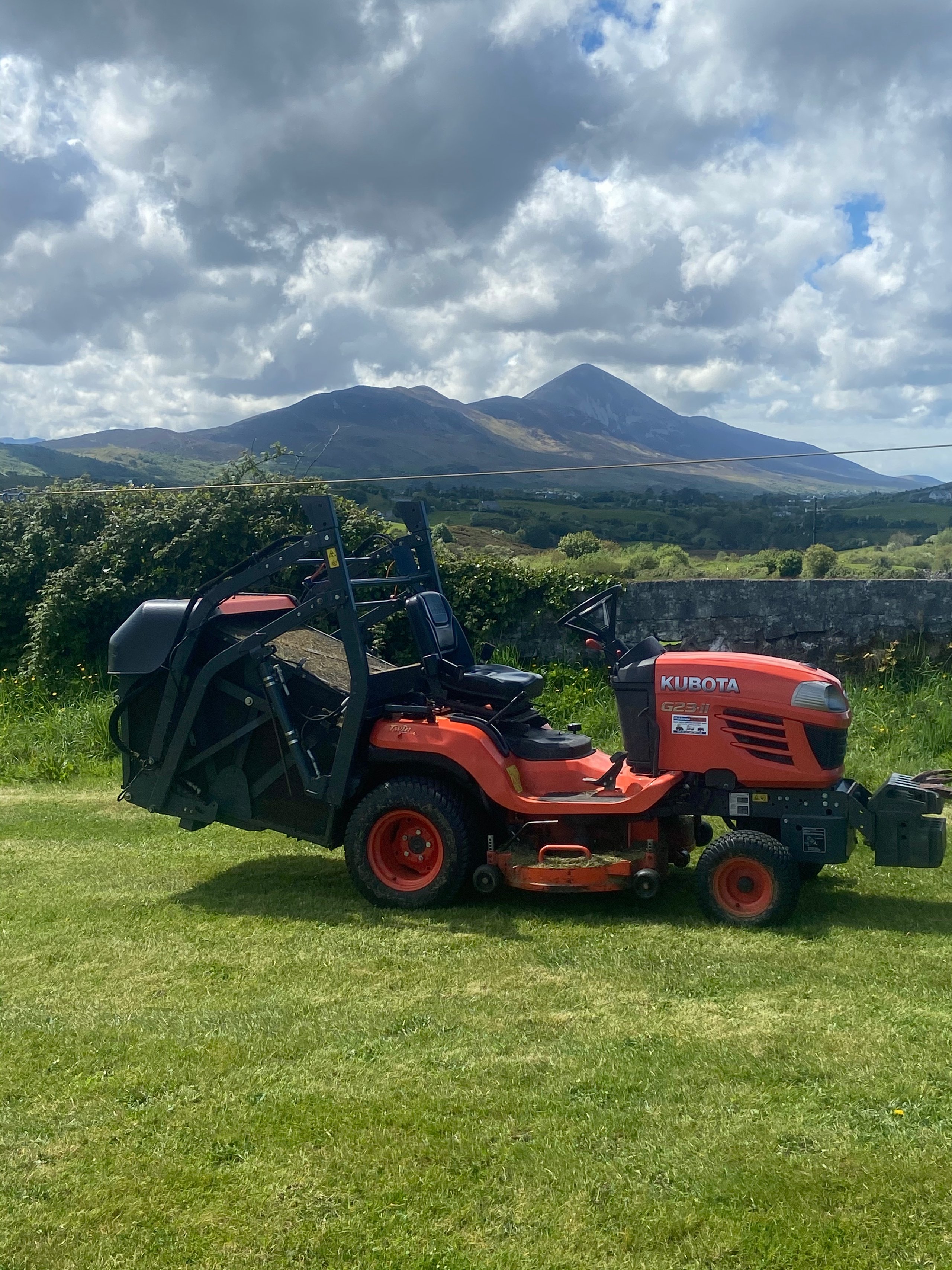 Orange Kubota G23-II ride-on lawn mower on green grass with a stone wall and mountains under a partly cloudy sky.