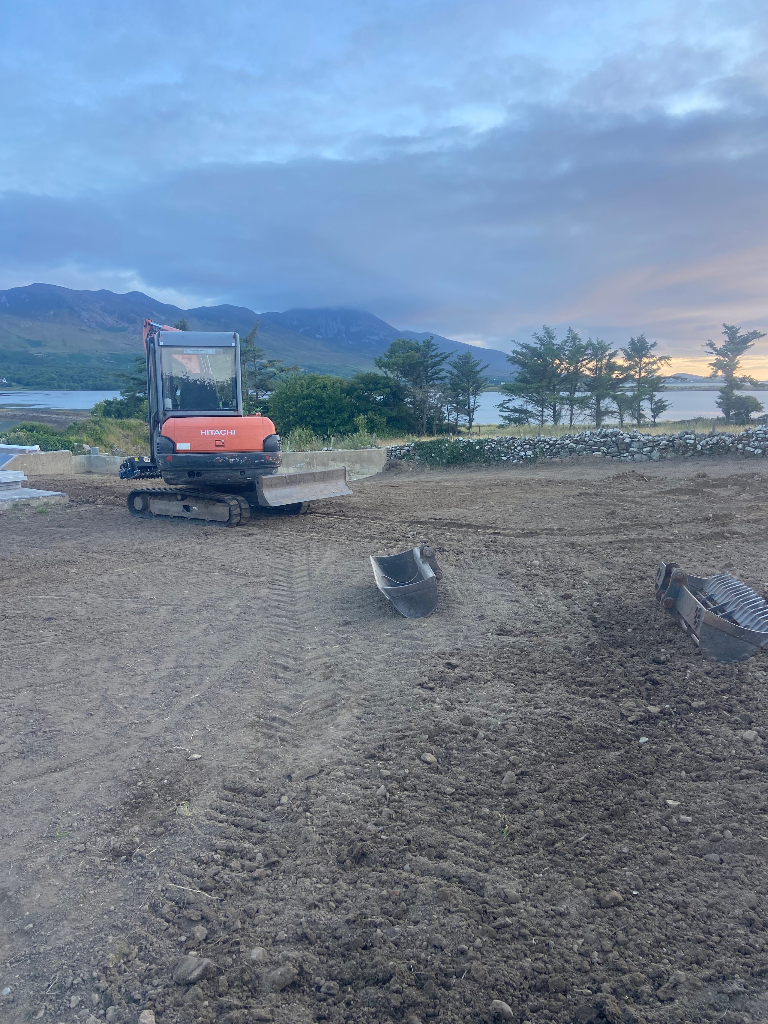 Orange Hitachi excavator on a cleared dirt construction site with mountains, trees, and a lake in the background at sunset.