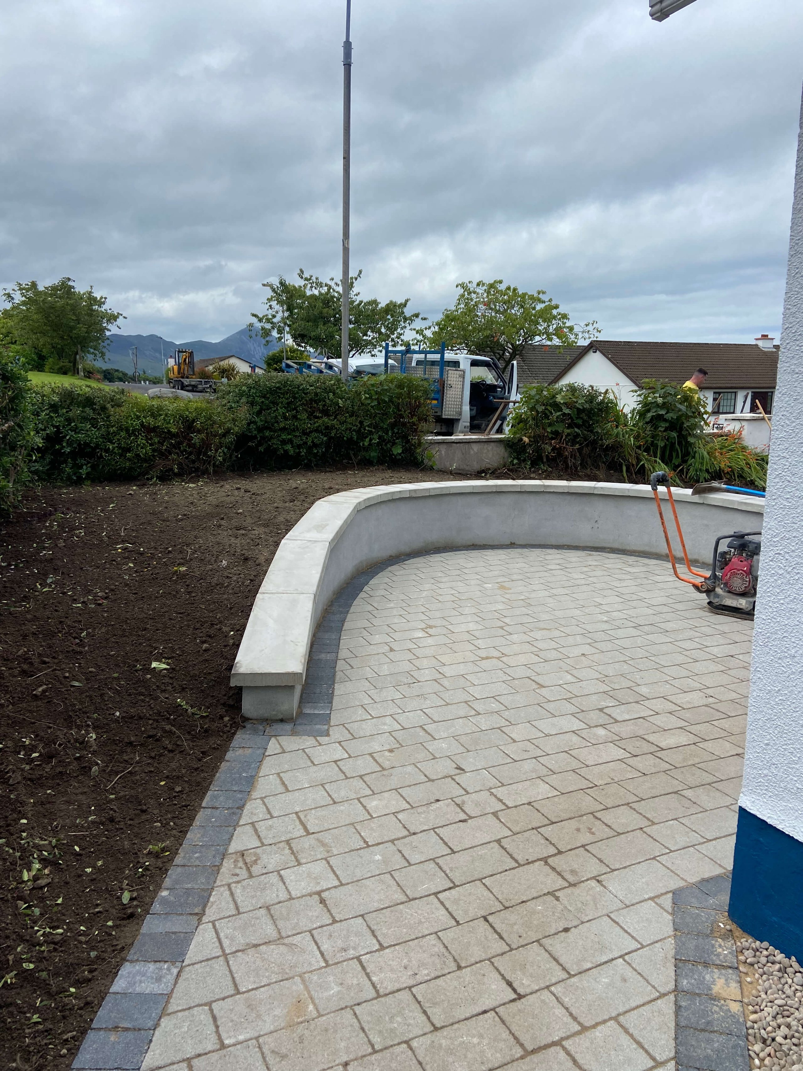 Curved concrete bench bordering a newly paved patio with a patch of freshly tilled soil and shrubbery under a cloudy sky.