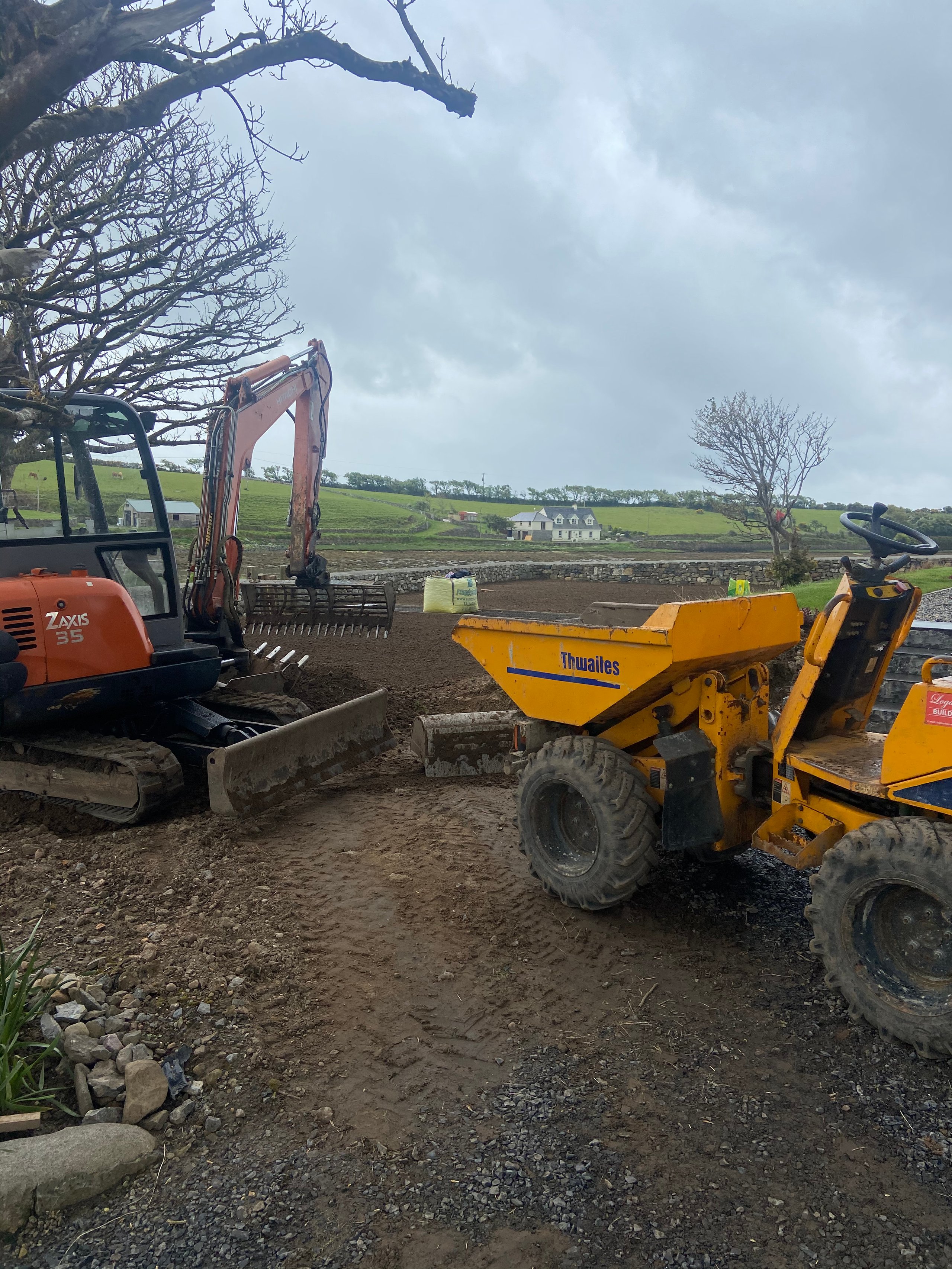 Construction site with a yellow Thwaites dumper and an orange Hitachi Zaxis 35 excavator on muddy ground near fields and a house.