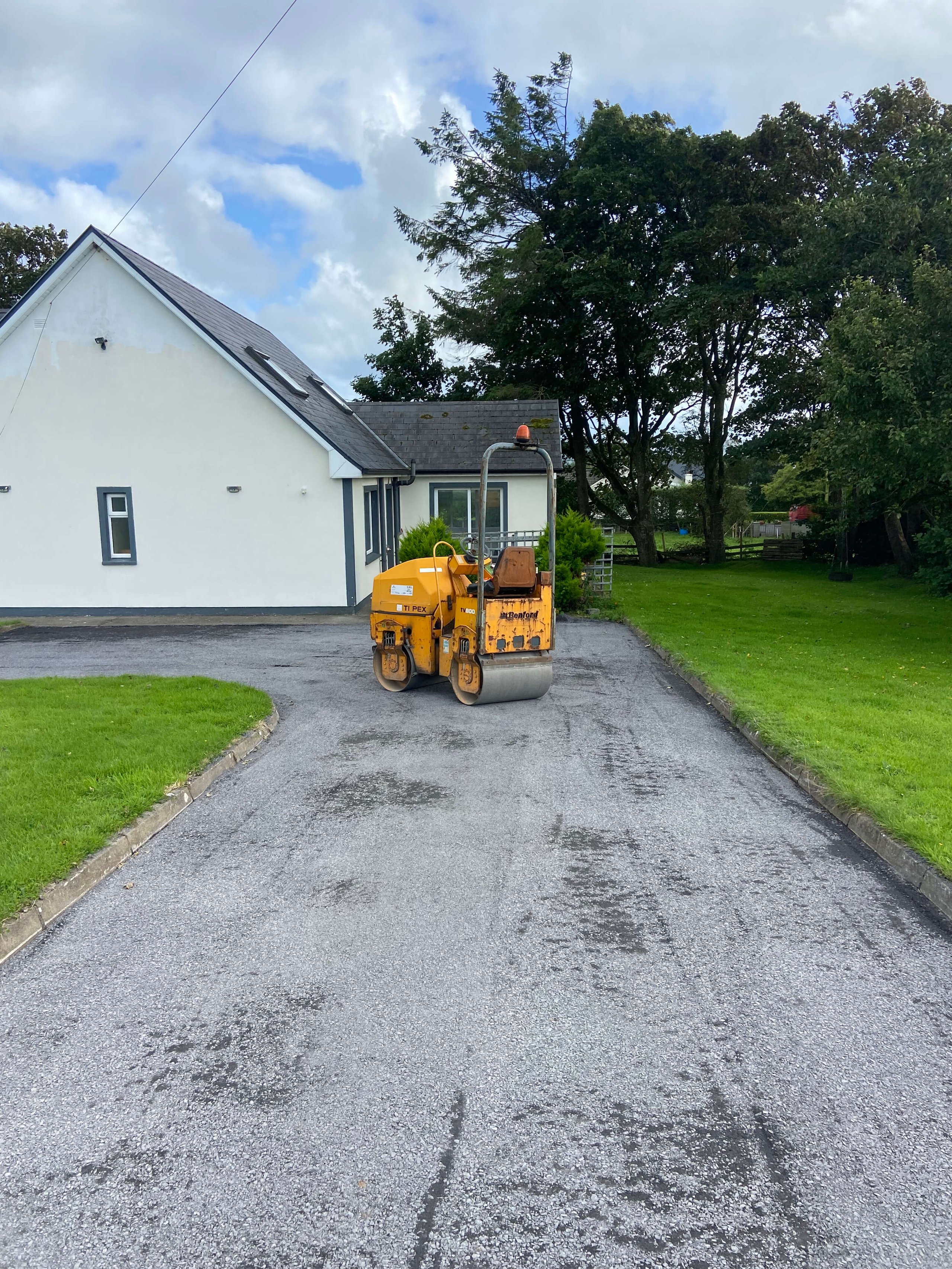 Yellow road roller parked on a freshly paved driveway beside a white house with green lawns and trees.