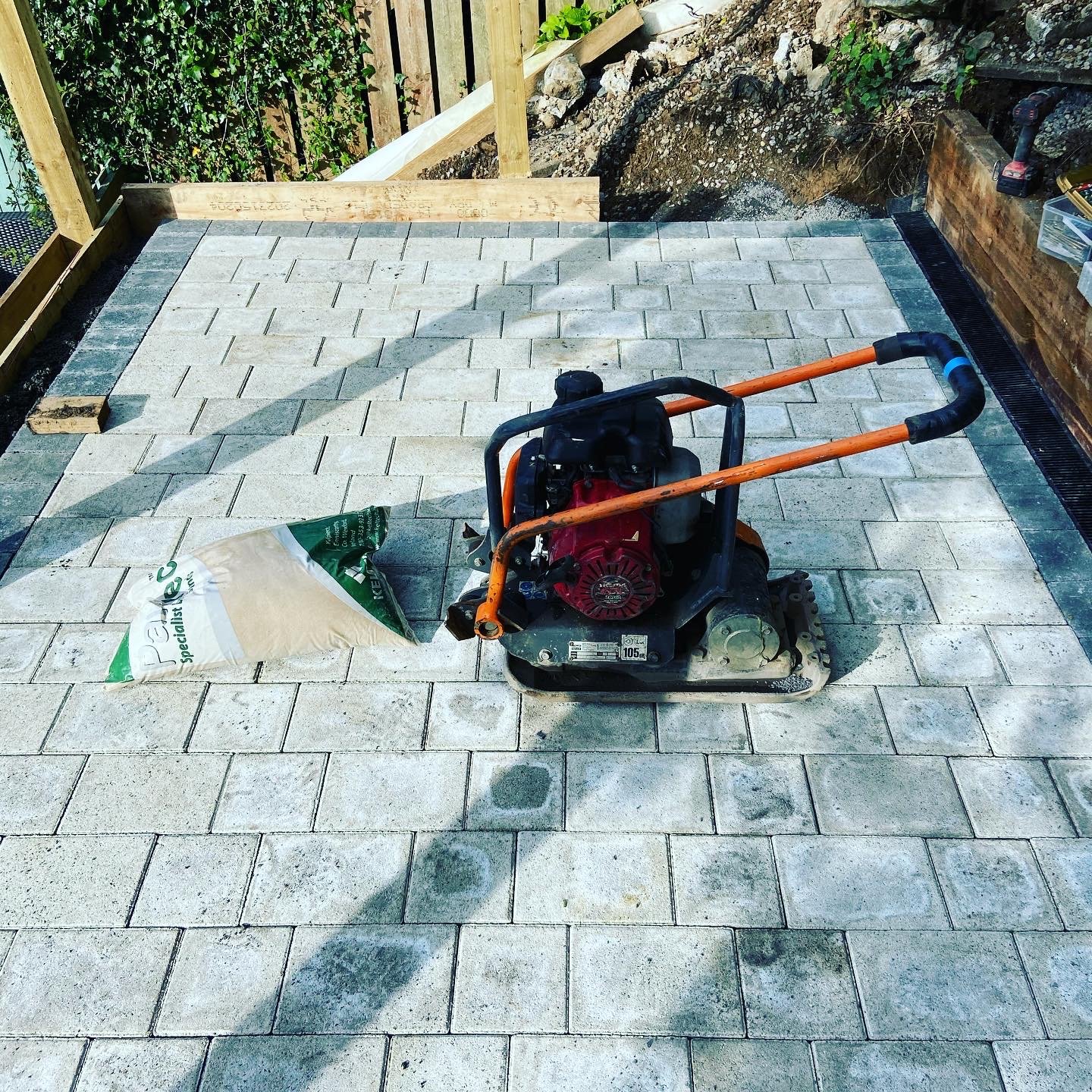 Outdoor paved area with a compactor machine and a partially opened bag of sand on gray pavers.