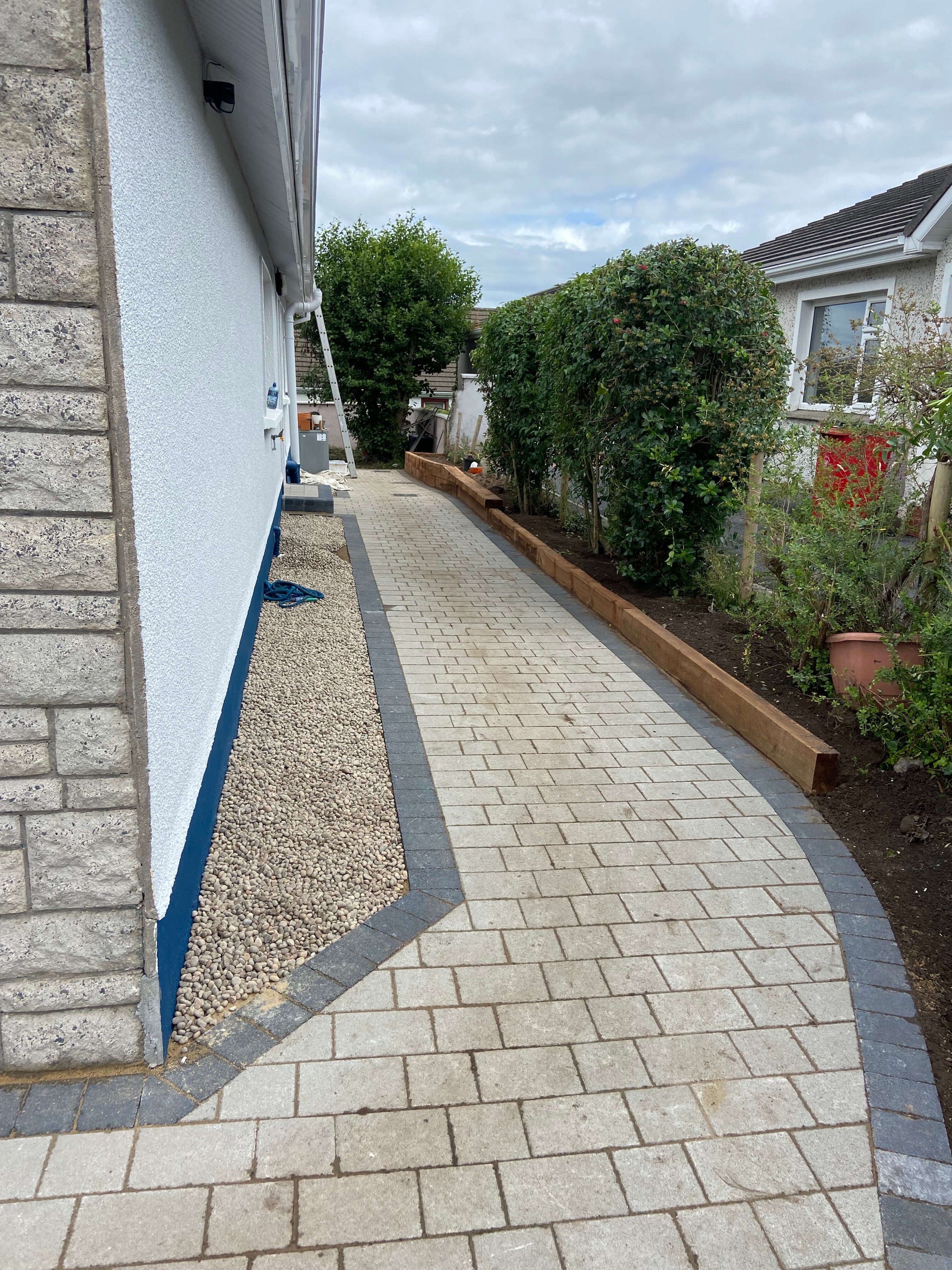Curved paved walkway bordered by a white building with stone accents on the left and a wooden-edged garden with bushes on the right.
