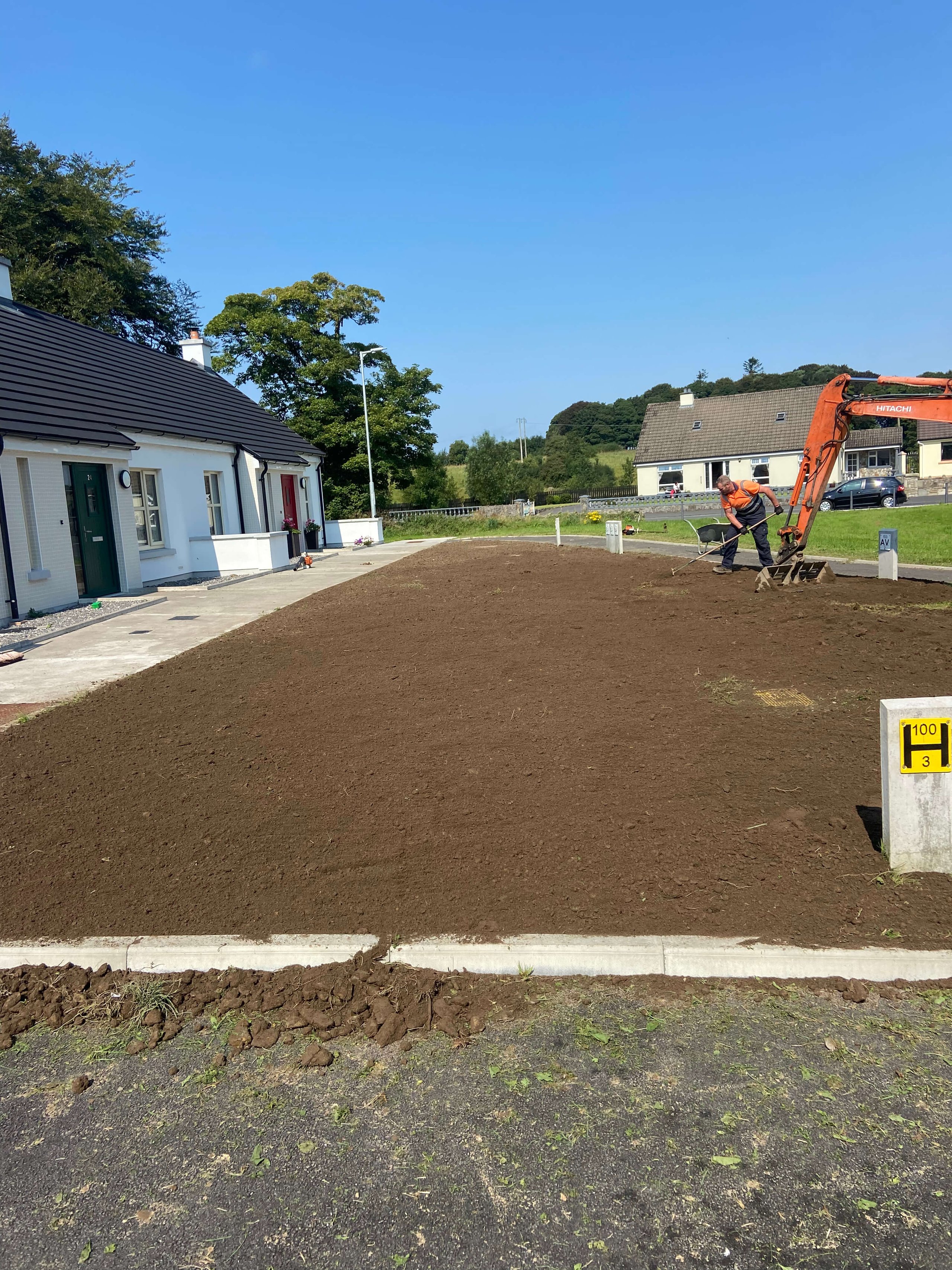 Man working with a rake next to an orange excavator on freshly tilled soil in front of white houses under a clear blue sky.